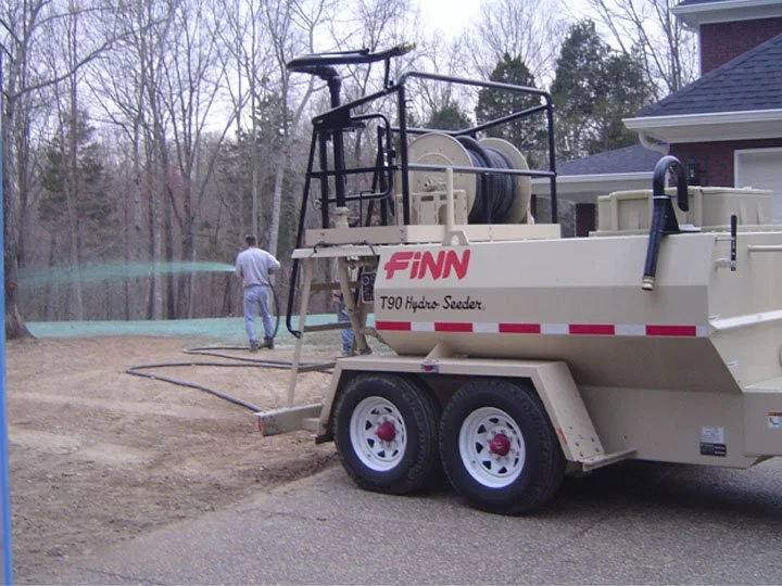 A worker in light-colored clothing operating a Finn T90 Hydro Seeder on a construction or landscaping site, with house and trees in the background.