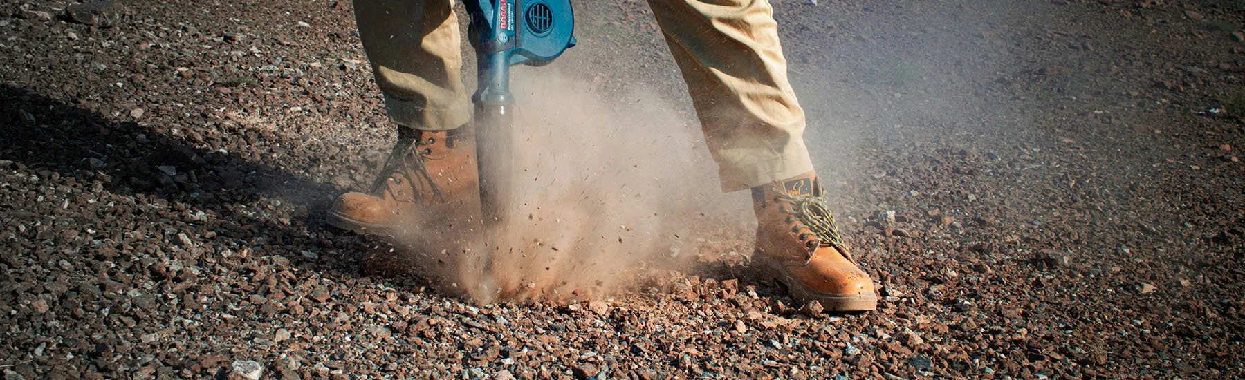 A worker in tan boots and beige pants using a blue pneumatic jackhammer on a gravel road, causing dust and small rocks to fly up.