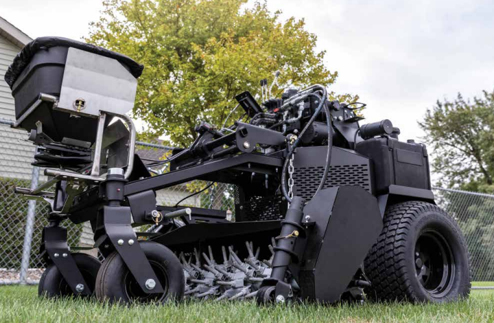Large, black, robotic-looking machine with wheels on a grassy field, surrounded by trees and a chain-link fence.