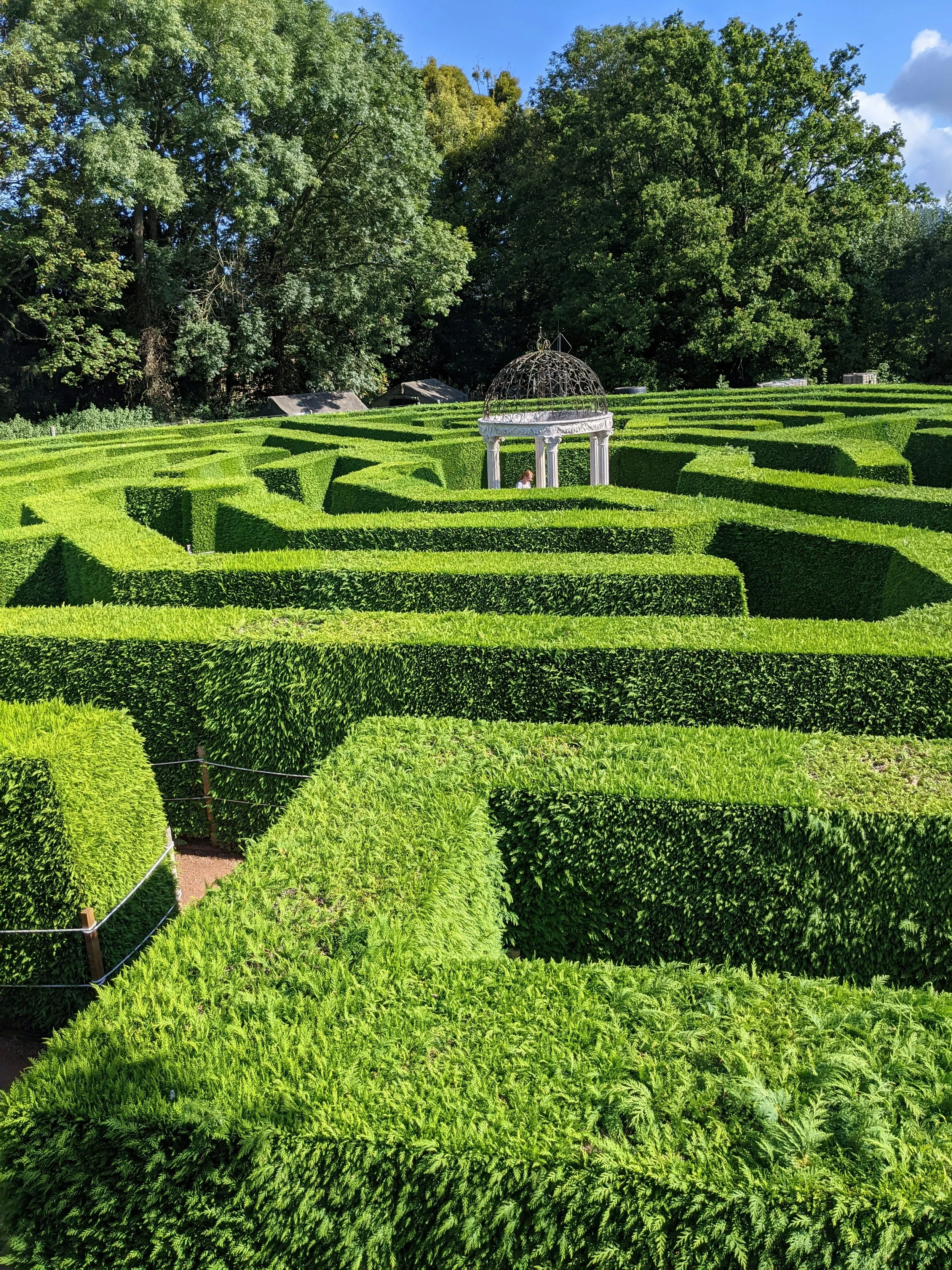 A lush green maze garden with neatly trimmed hedges surrounds a white classical gazebo with a domed top. Tall trees and a blue sky with some clouds are in the background.