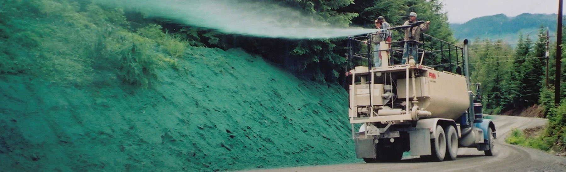 A truck equipped with a sprayer attached to the back is spraying green liquid on a hillside along a rural road. Two workers are on top of the truck operating the equipment.