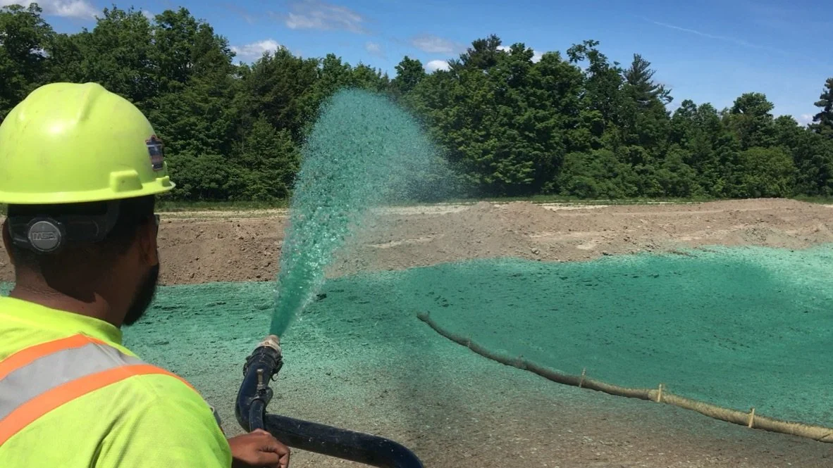 A construction worker wearing a yellow helmet and high-visibility vest spraying a green-colored material onto the ground in an outdoor setting with trees and a clear blue sky.
