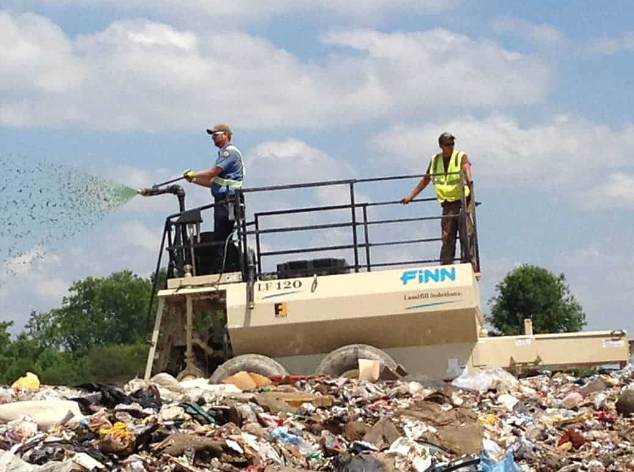 Two workers in safety vests standing on a vehicle at a landfill, one spraying water to control dust, with piles of trash in the foreground and a partly cloudy sky.