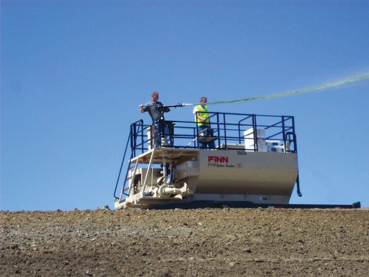 Two people standing on a blue safety platform attached to a white rail-mounted work vehicle on a gravel surface, spraying a substance into the sky during daytime.
