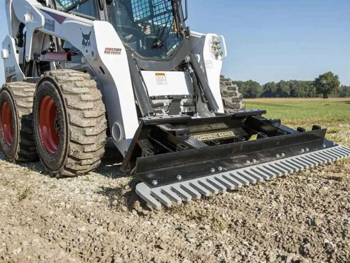 A skid steer loader with a black grapple attachment on rocky ground in an open field under a clear sky.