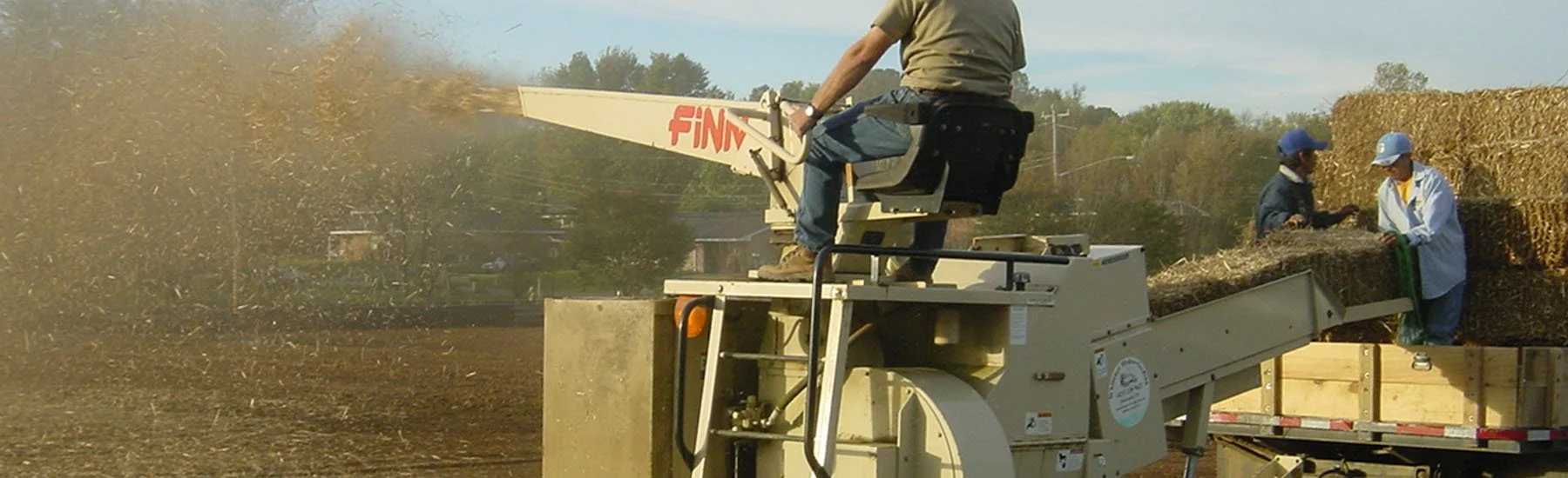 Two workers unload hay bales onto a trailer while a third person operates a hay baler machine labeled 'FINN' in a rural farm setting.