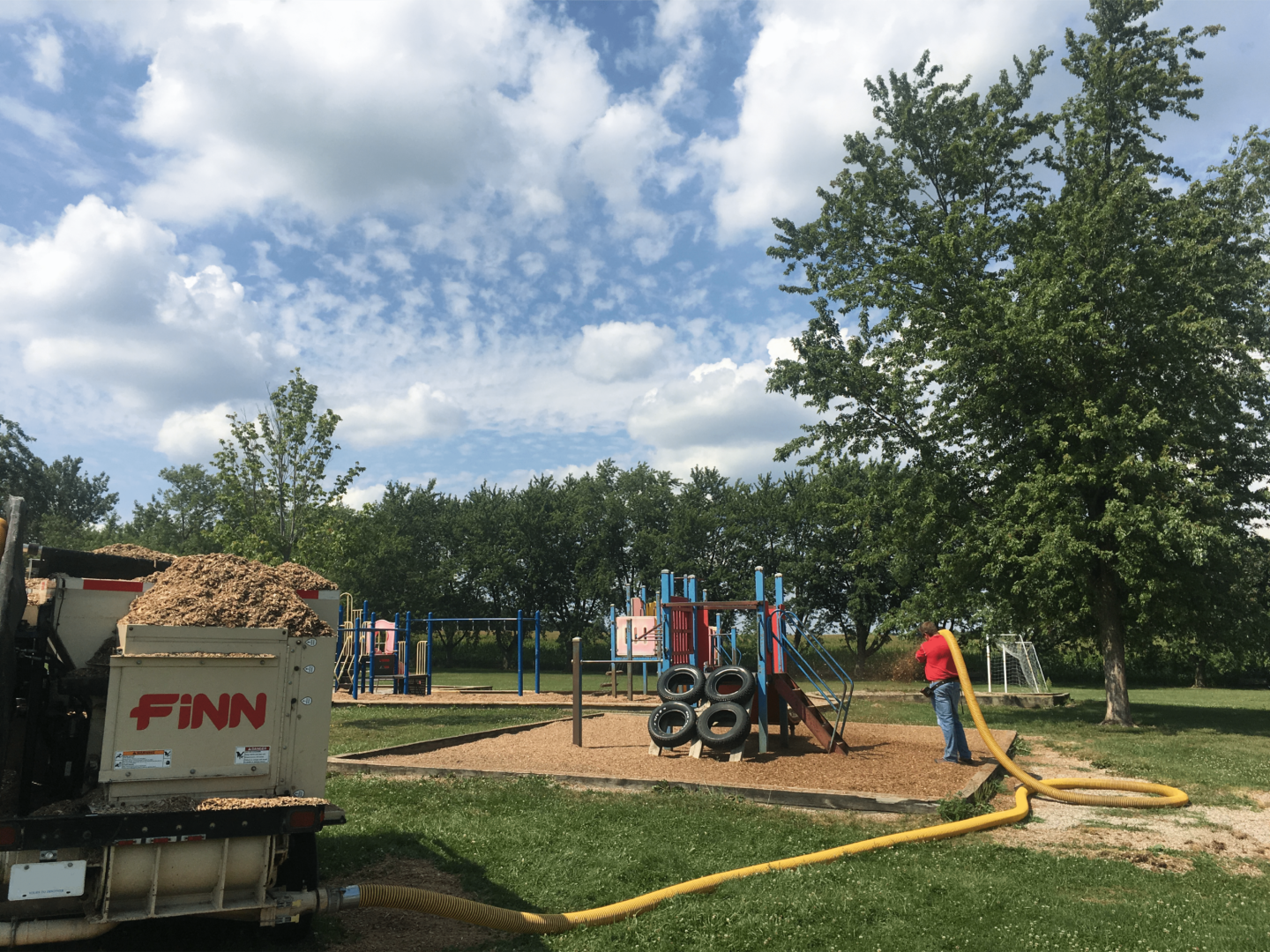 A playground with a slide, climbing structures, and tires, with a man handling a large yellow hose connected to a truck involved in maintenance work, and trees and blue sky with some clouds in the background.