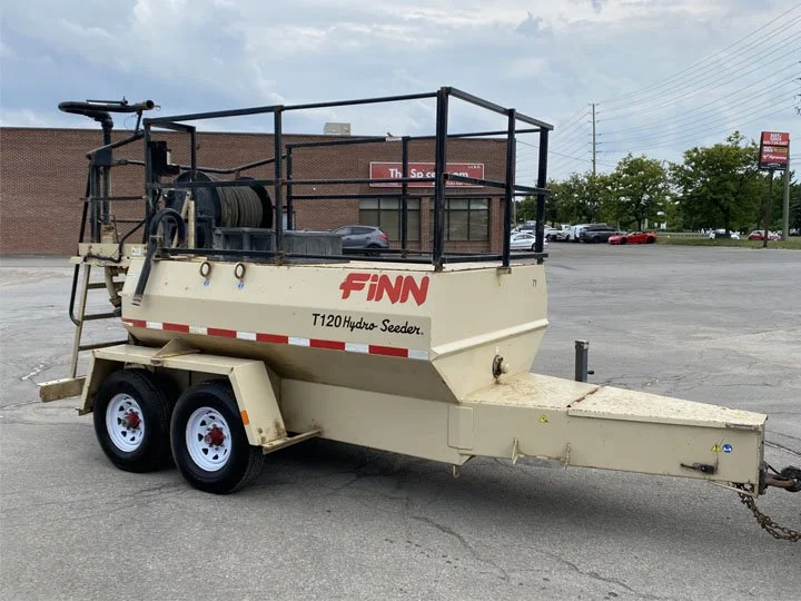 A Finn T120 Hydro Seeder trailer with a hose reel on top, parked in an empty parking lot outside a commercial building.
