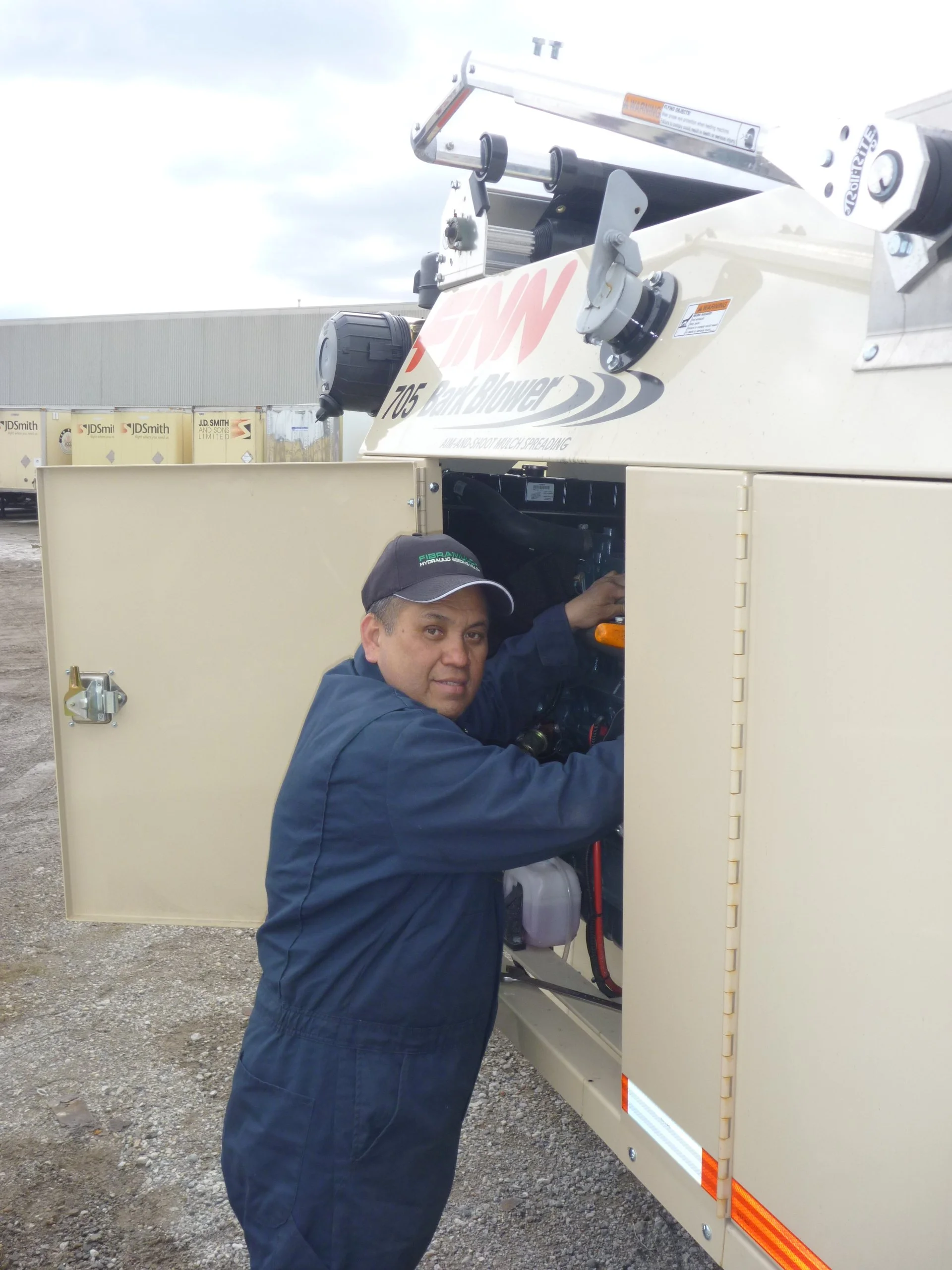 A man in a blue jacket and black cap working inside a white utility trailer with the label 'Xtreme Bed Blower' and the words 'Airless Spray Technology Spreading' on it, outdoors on a cloudy day.
