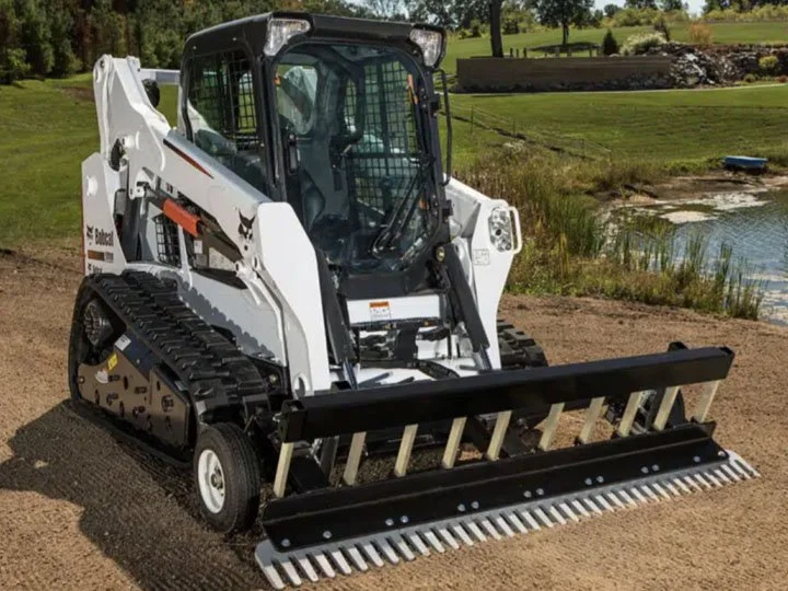 A compact white and black skid steer loader with a front-mounted rake attachment on a construction site near a pond with grass and trees in the background.