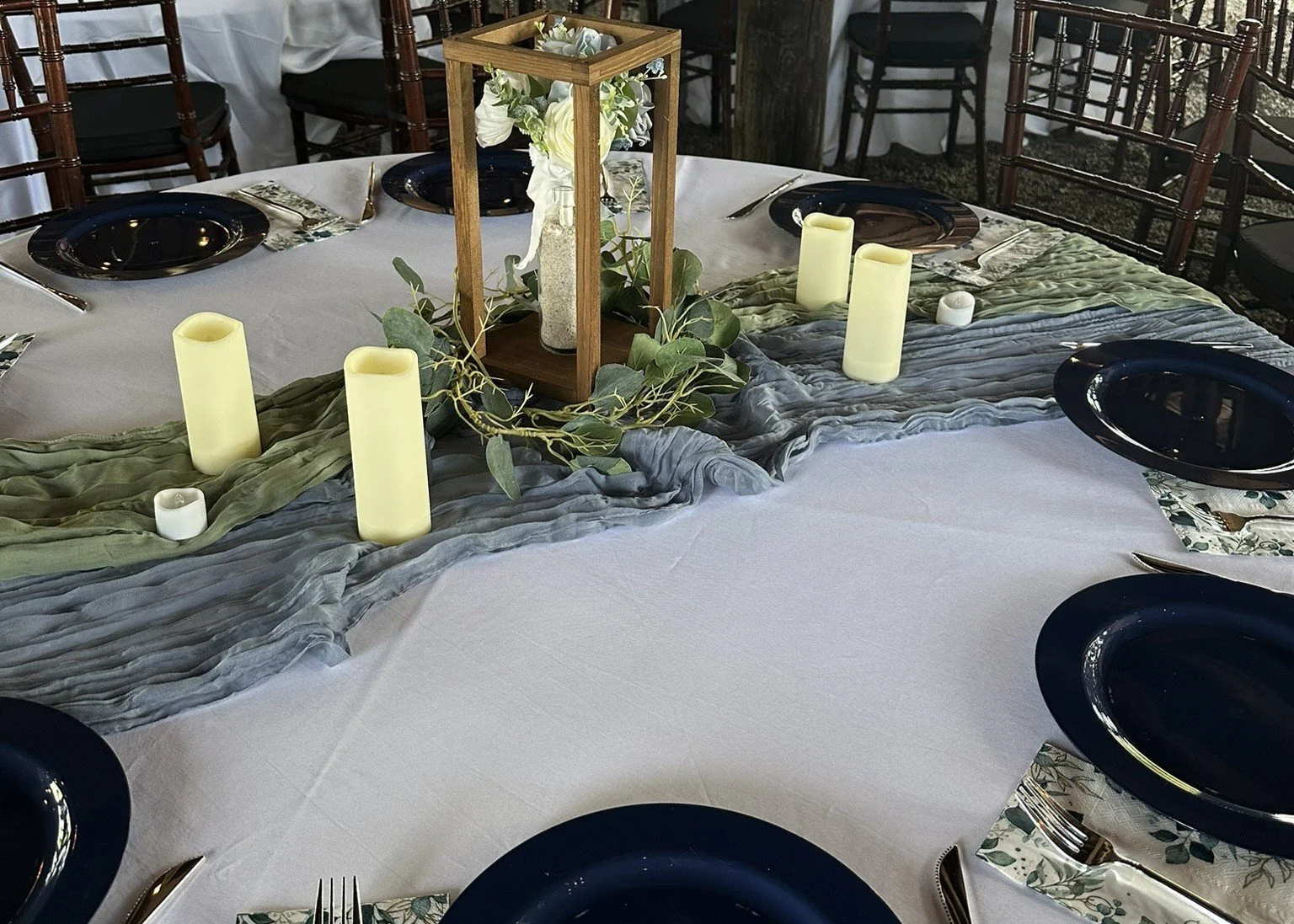 Round banquet table with a white tablecloth and a gray and green draped runner. Centerpiece features a glass vase with white flowers inside a wooden frame, surrounded by greenery and candles in yellow and white. Black plates, silverware, and floral-patterned napkins are set around the table, with wooden chairs in the background.