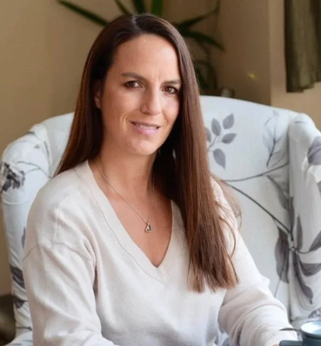 A woman with long brown hair sitting on a floral-patterned chair in a cozy room.