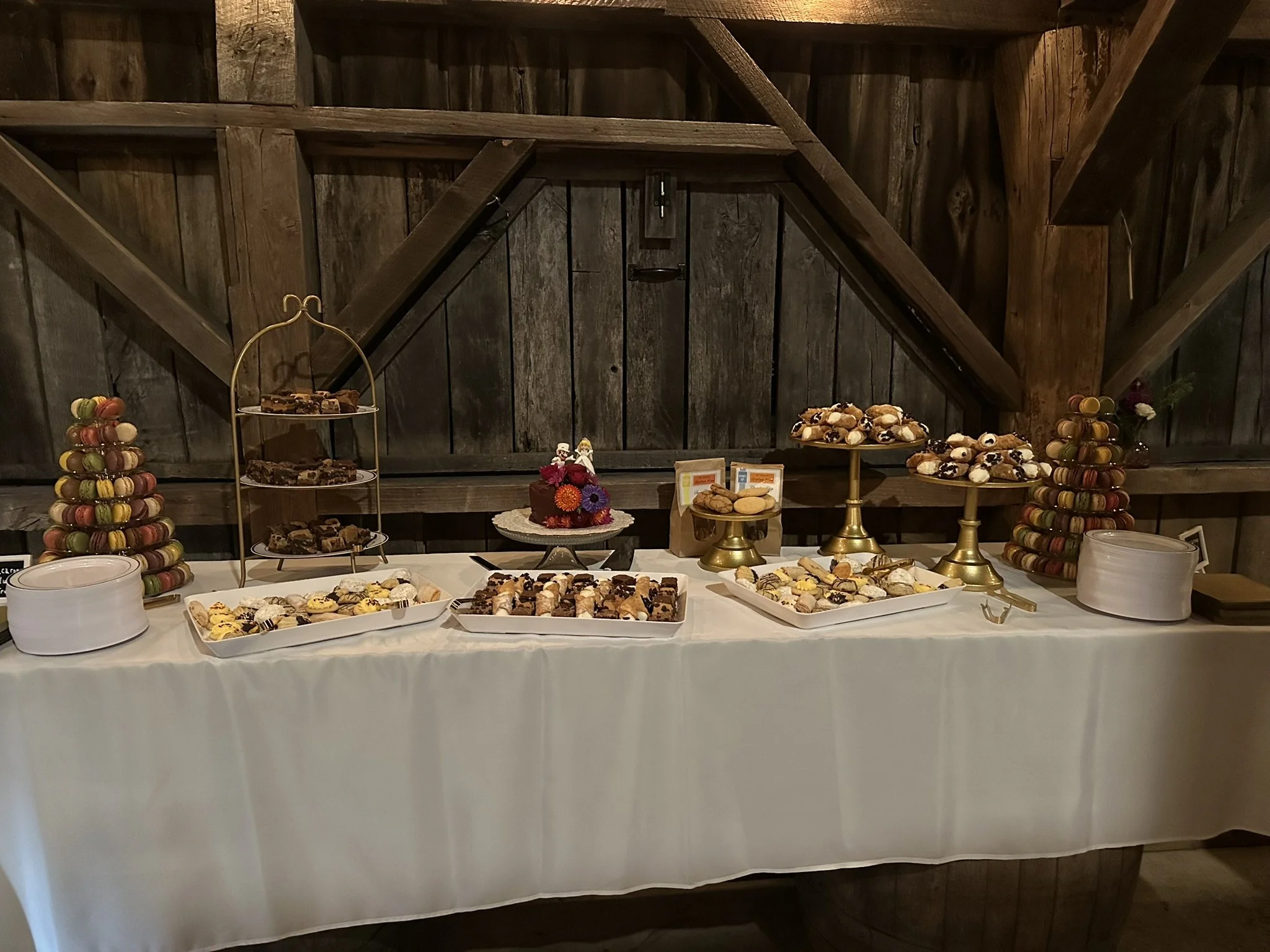 A dessert table set up against a rustic wooden wall with an assortment of treats, including cookies, pastries, and macarons displayed on white plates and gold tiered stands.