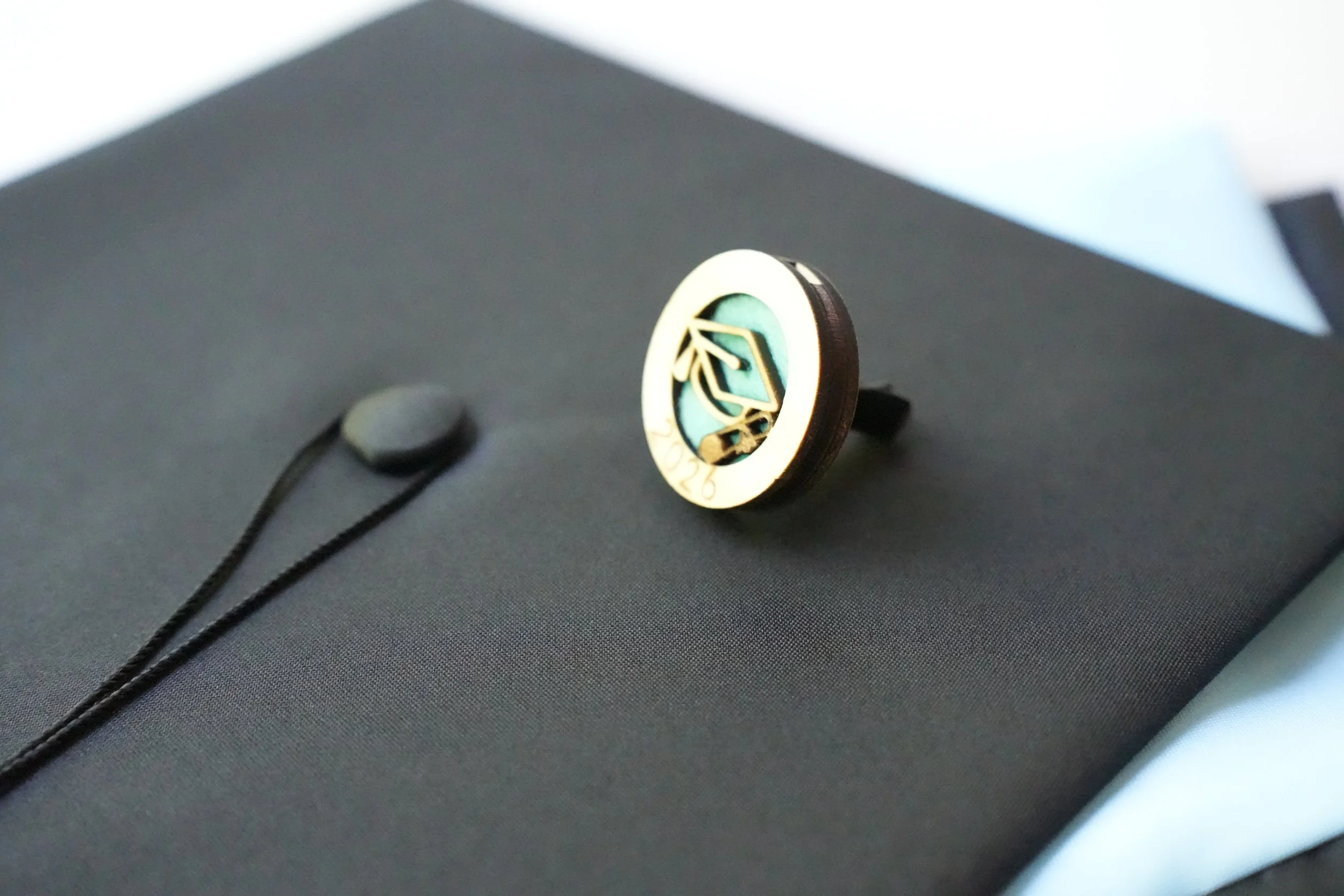 Graduation cap car freshener design with a personalized circular emblem and black ribbon.