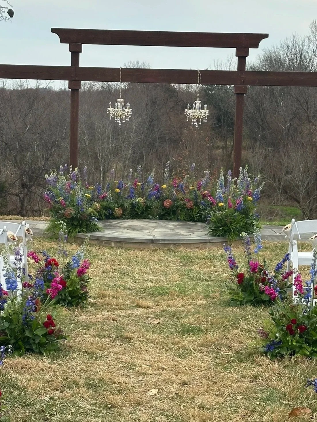 Outdoor wedding or event setup with a wooden arch, two hanging chandeliers, and floral arrangements with purple, pink, and red flowers on a grassy area in front of wooded trees.