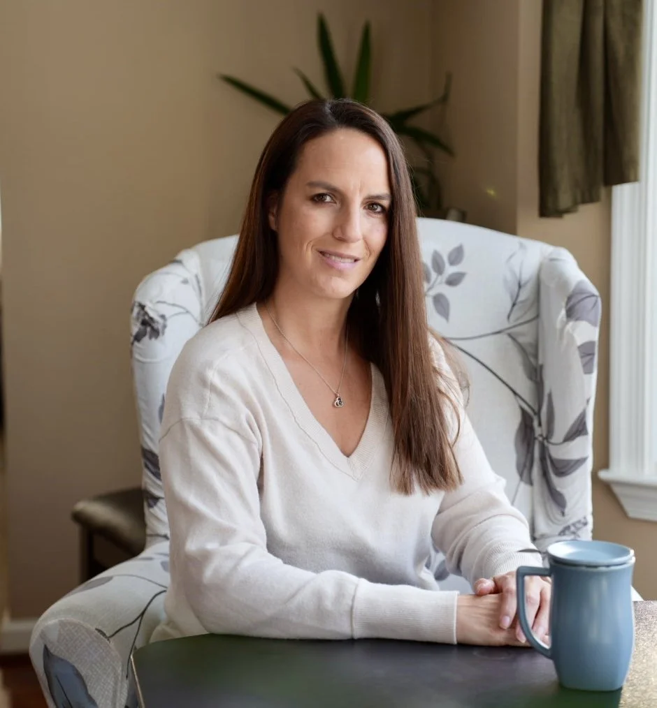 A woman with long brown hair and a white sweater sitting at a table with a blue mug, in a cozy room with a floral armchair, a plant, and window with curtains.