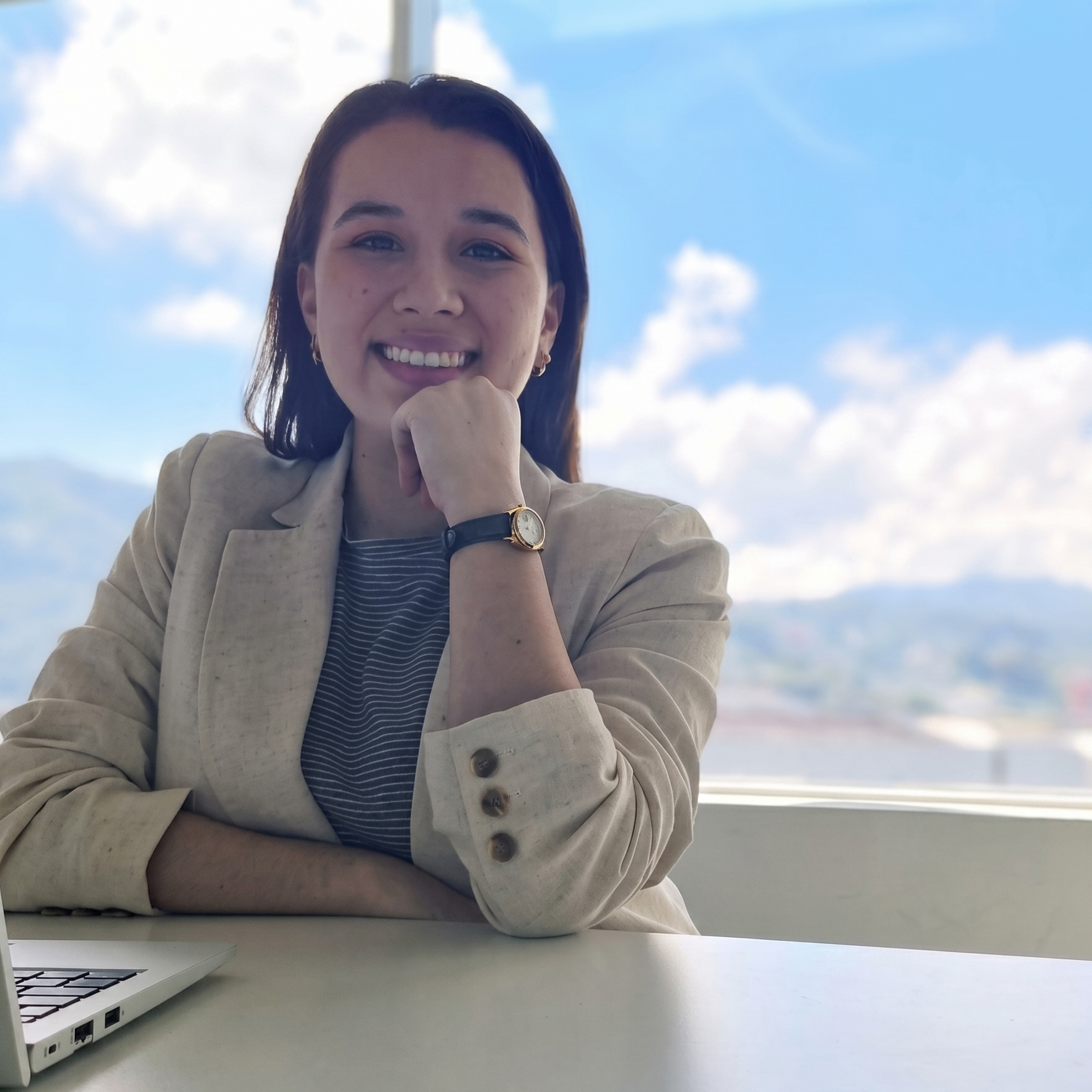 A woman with shoulder-length dark hair and a beige blazer smiling, resting her chin on her fist, sitting at a desk with a laptop, with a bright sky visible through a window behind her.