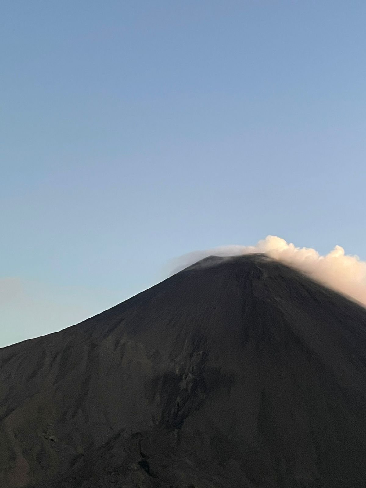 A photograph of a volcanic mountain with a plume of smoke and ash coming from its summit, set against a clear blue sky.