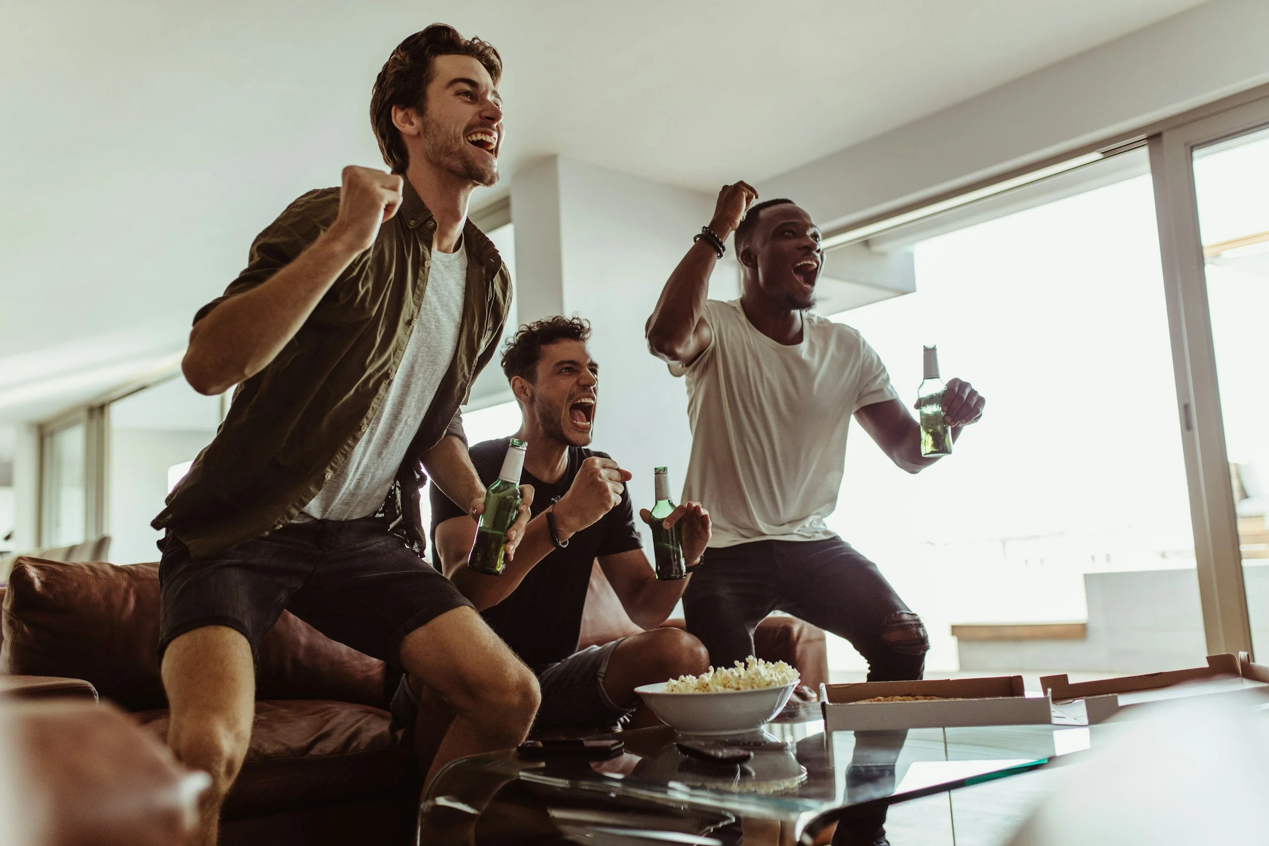Three young men watching football world cup 2026 games on TV, cheering, holding beer bottles, and excitedly celebrating in a living room.