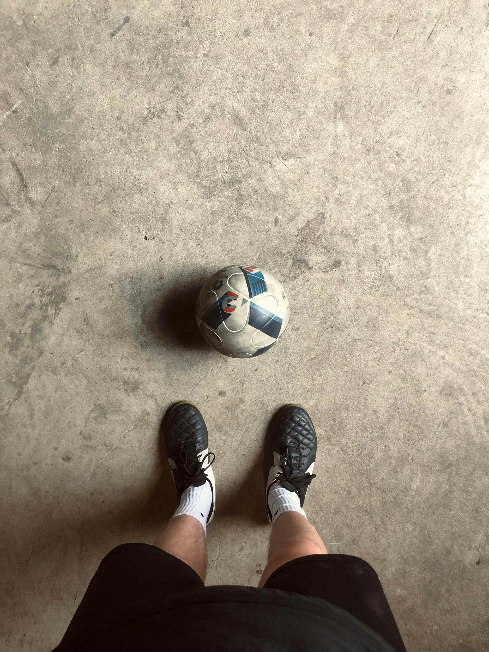 A person standing on a concrete floor, wearing black shorts, white socks, and black athletic shoes, with a football in front of them. The person is playing football solo.