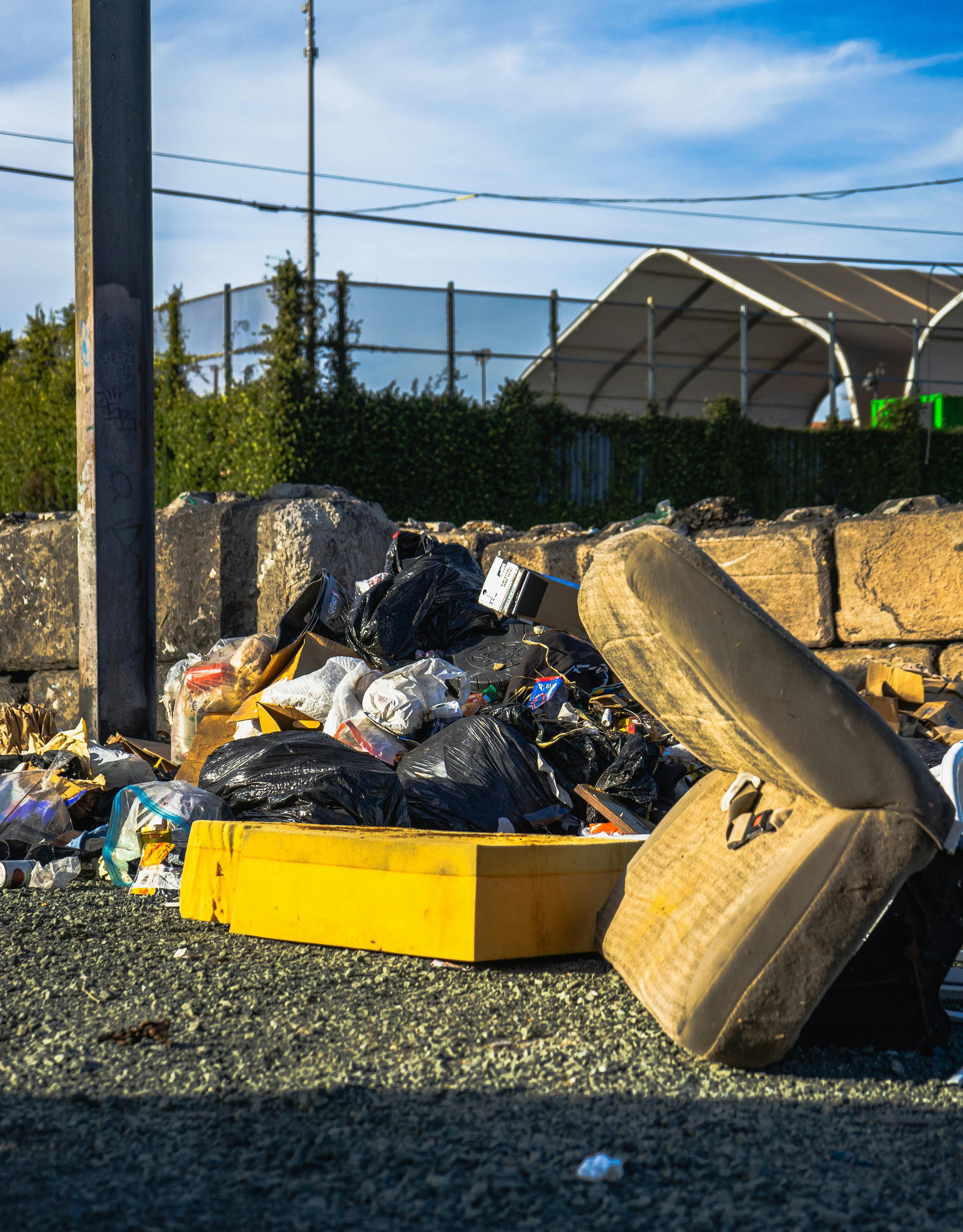 A pile of trash including black garbage bags, a yellow dumpster, and discarded debris on asphalt ground, with a pole and a fence with trees and a structure in the background.