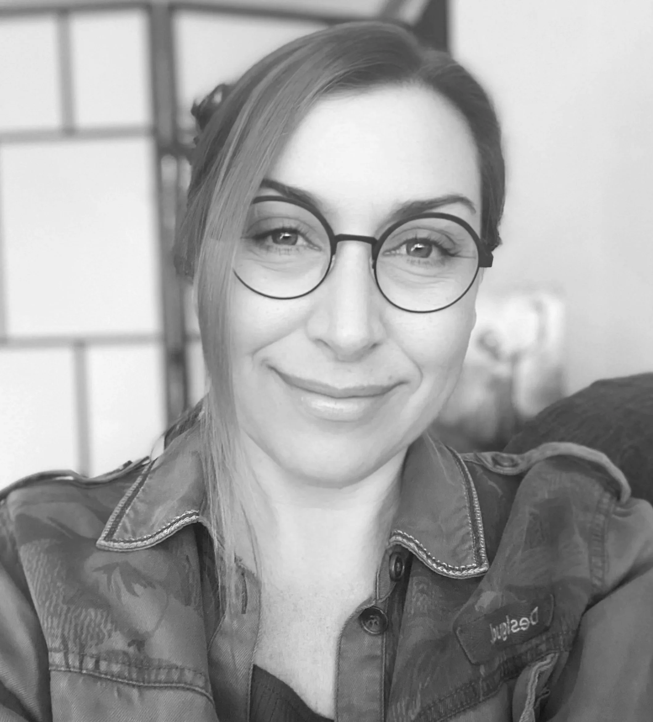 A black-and-white photograph of Lisa Greenaway. She is wearing round glasses, with straight hair parted to one side, smiling slightly, dressed in a denim jacket, with a shoji screen in the background.