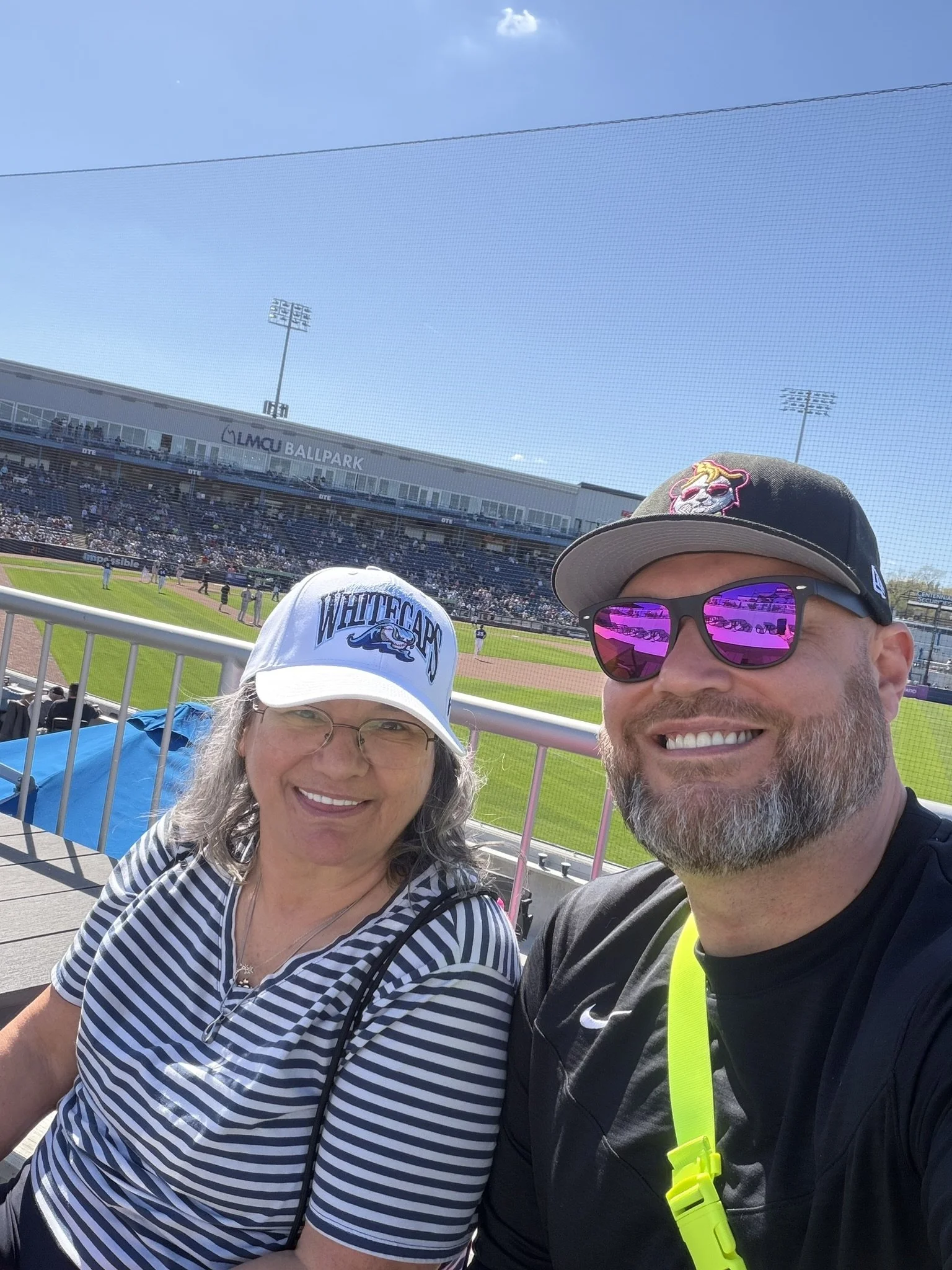 A mother and son taking a selfie at a baseball stadium on a sunny day, with the field and bleachers visible in the background.