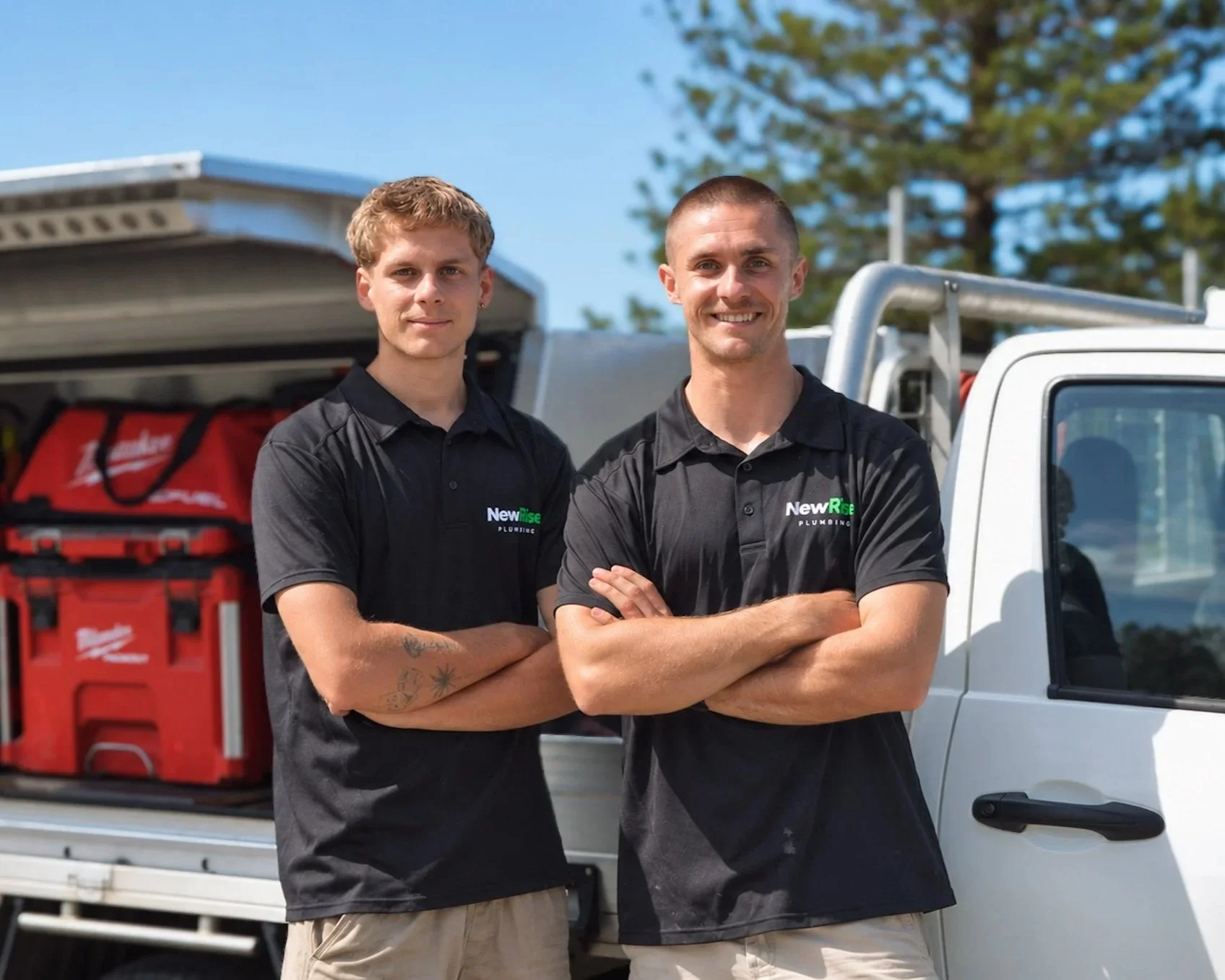 Jack and Cooper from NewRise Plumbing standing beside their fully equipped plumbing ute in Sydney. The truck's bed is open, showing red tools and equipment inside. Bright and sunny outdoor setting with a tree in the background.