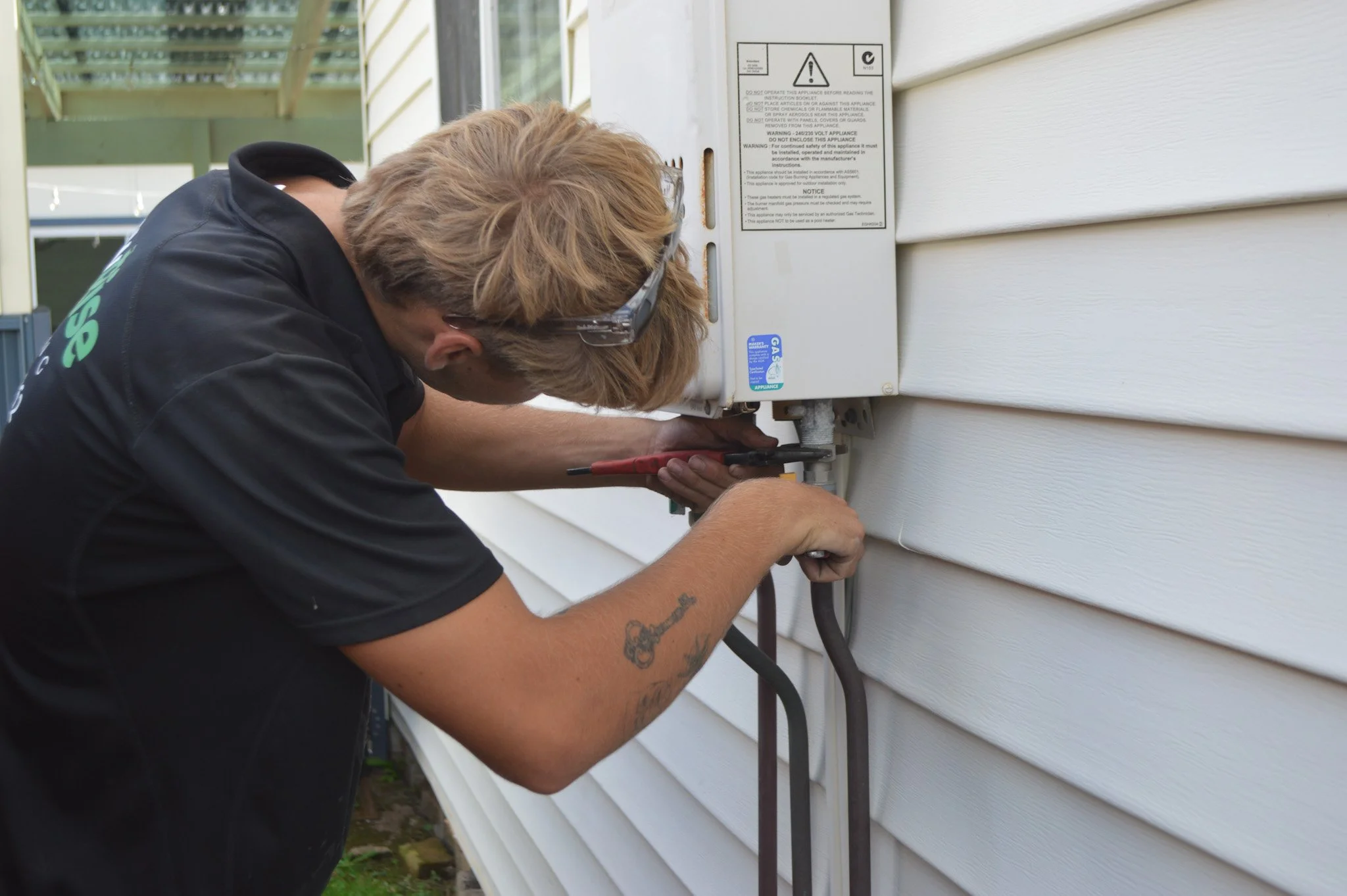 Cooper from Newrise Plumbing working on an outdoor electrical box attached to the side of a house, using a screwdriver, wearing safety goggles and Newrise Plumbing uniform.