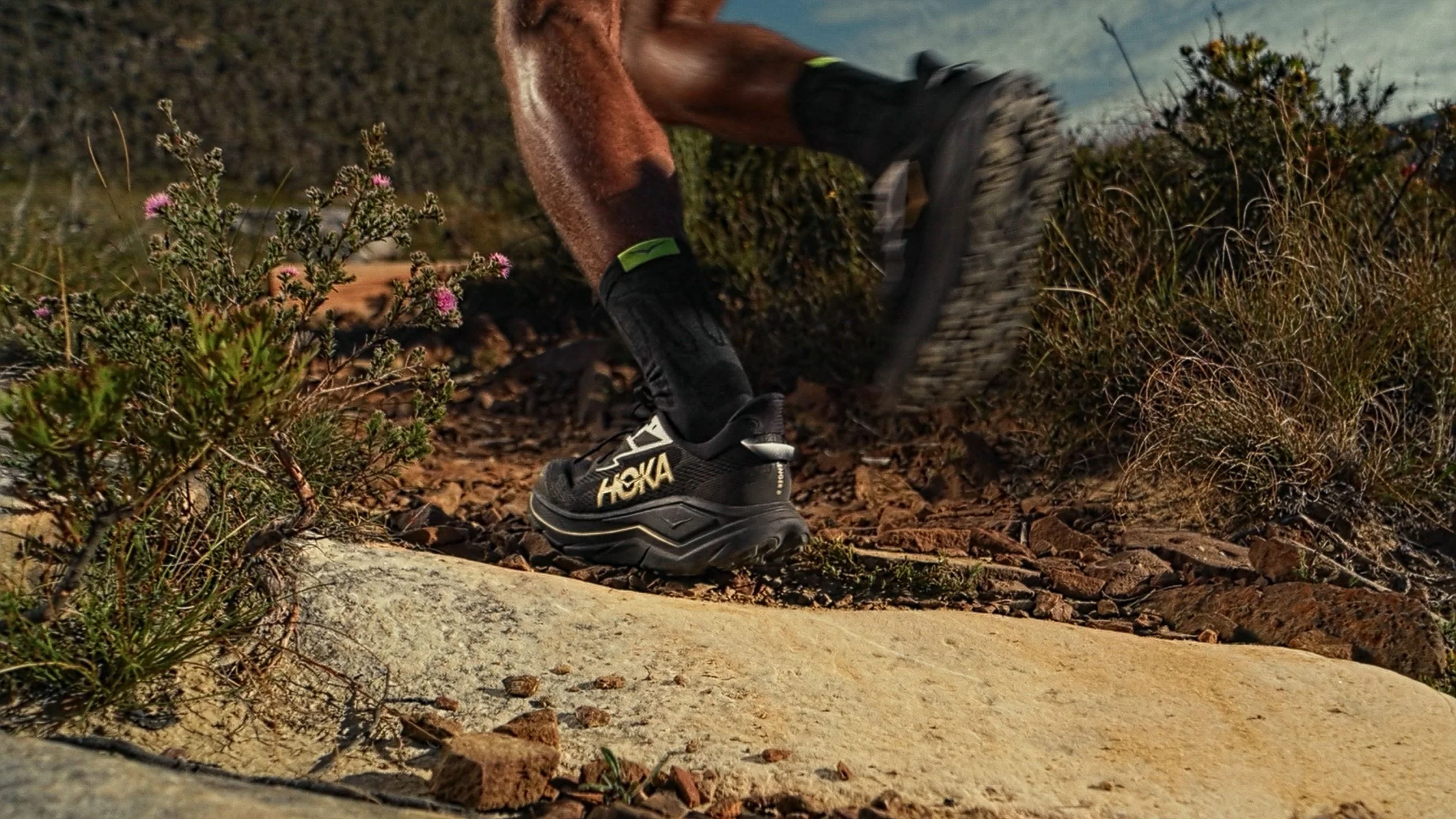 Close-up of a person's feet wearing black Hoka running shoes and black socks, walking on a rocky trail surrounded by desert plants and shrubs under cloudy sky.