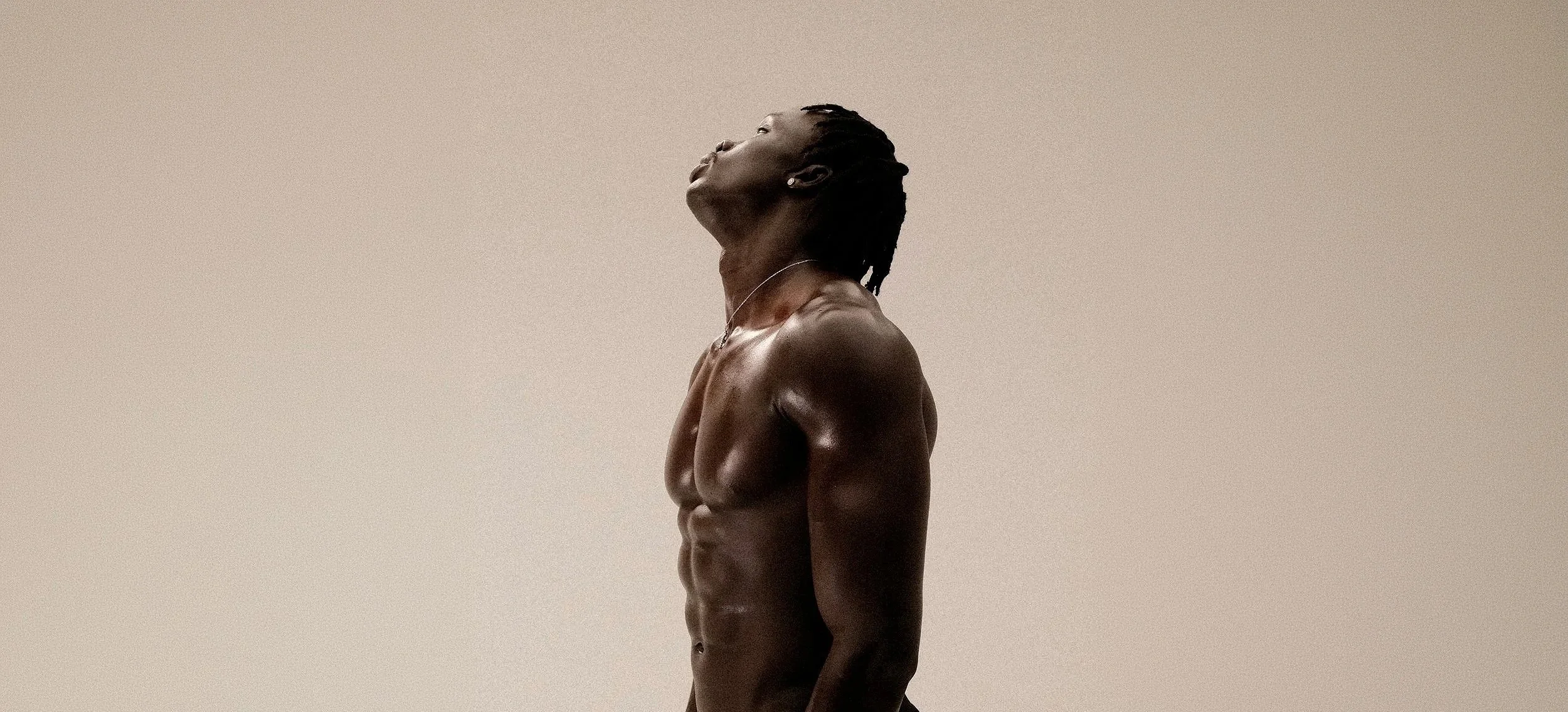 Side view of a shirtless black woman with short dreadlocks, standing against a neutral background with her head tilted back and eyes closed.