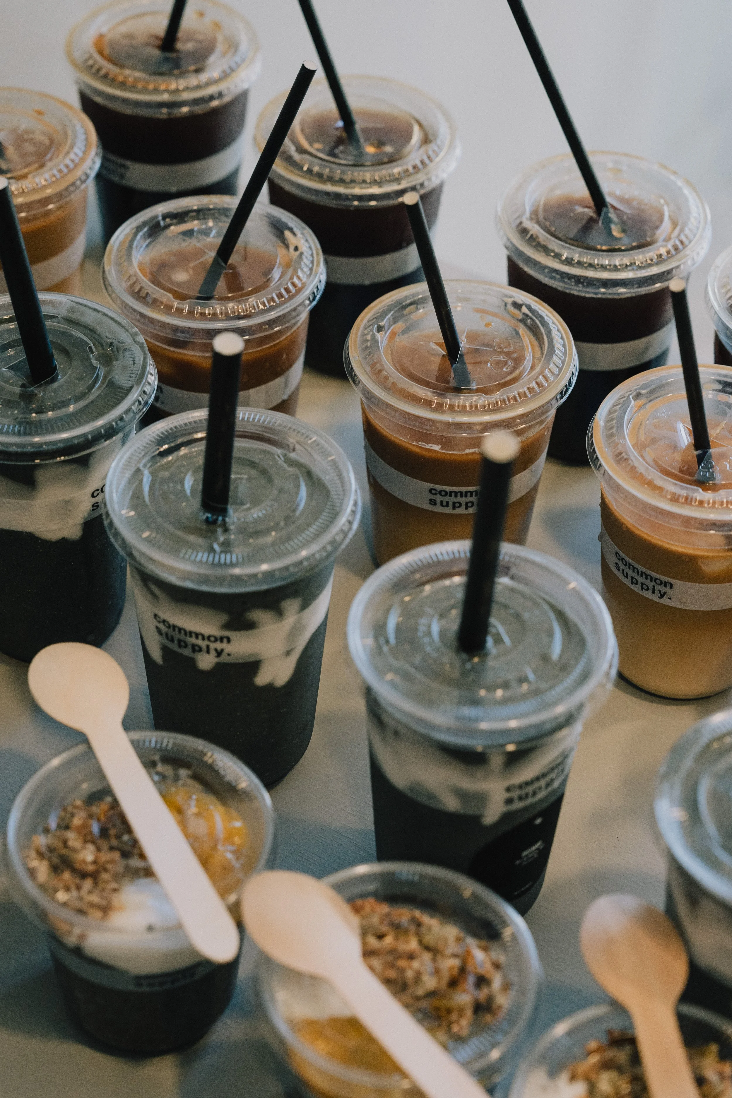 Multiple small plastic cups of iced coffee with black straws on a table. Some cups have a white band with black text reading 'common supply.' The beverages have varying degrees of cream and are topped with different toppings or toppings.