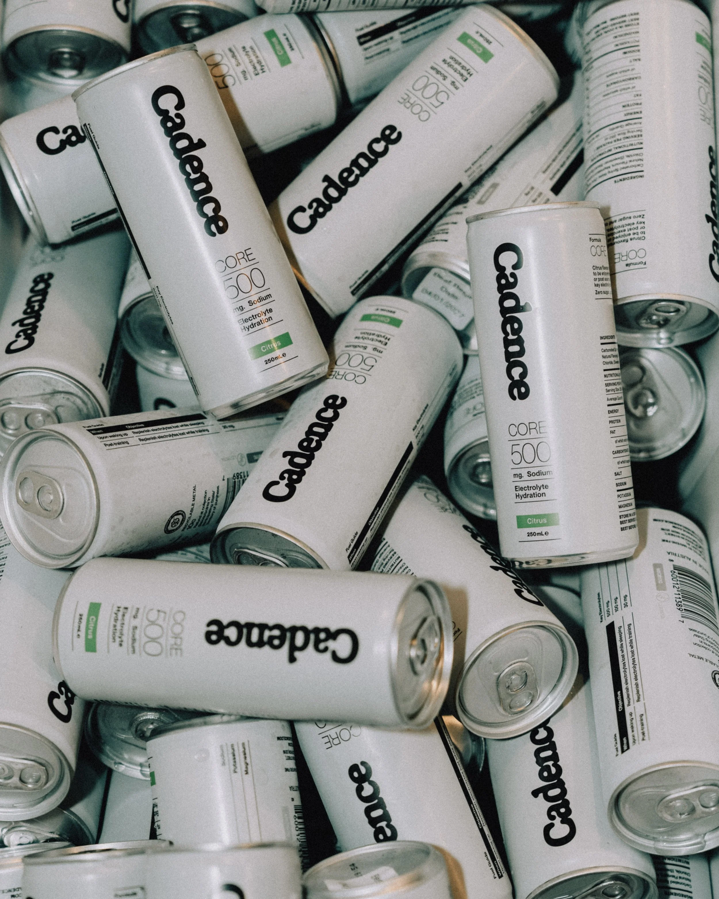 A pile of canned electrolyte drinks labeled 'Cadence,' with the flavor 'Citrus' and a dosage of '500 mg' sodium. The cans are silver with black and green text.