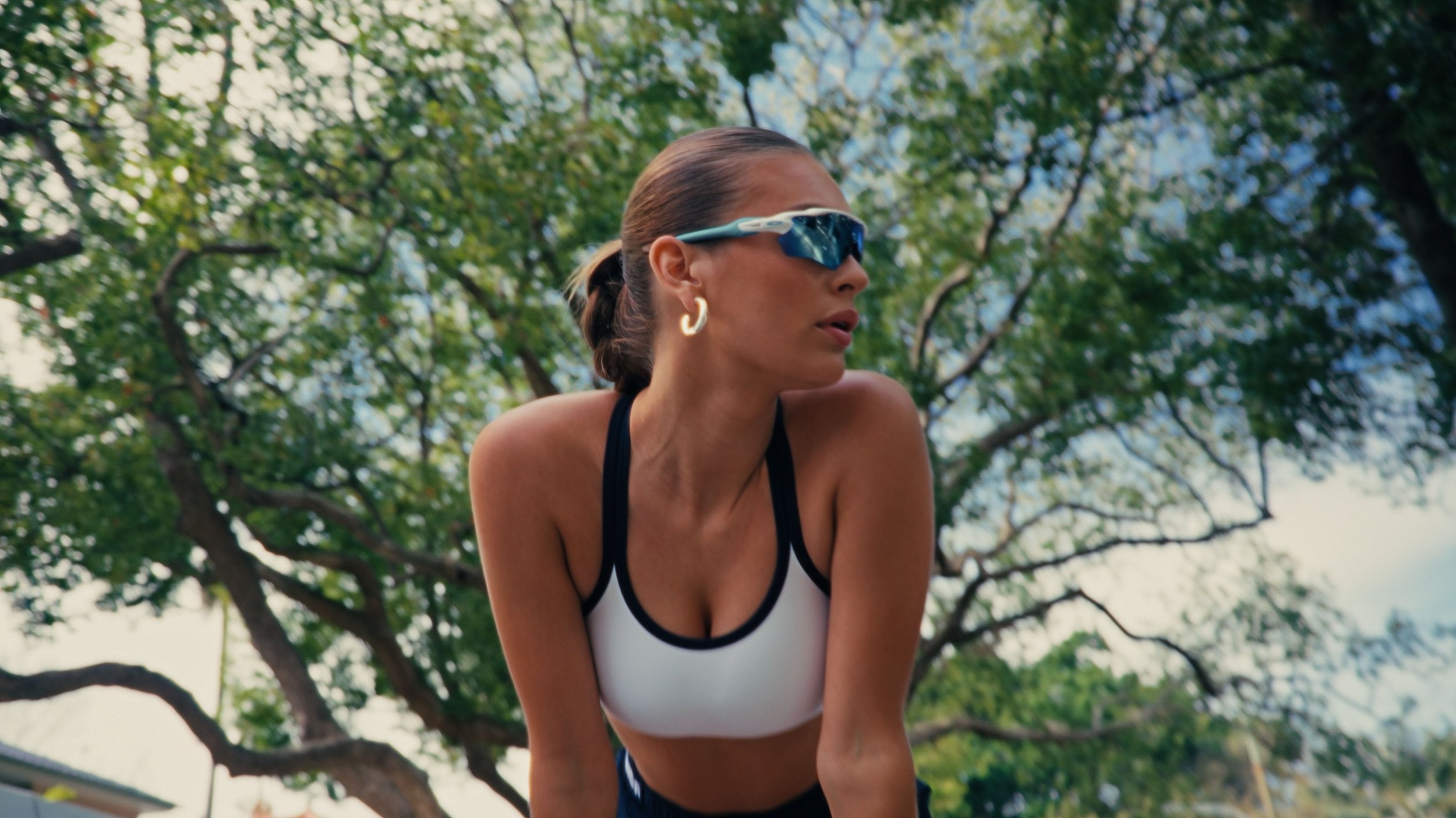 A woman wearing sports sunglasses and a sports bra, outdoors with green trees in the background.