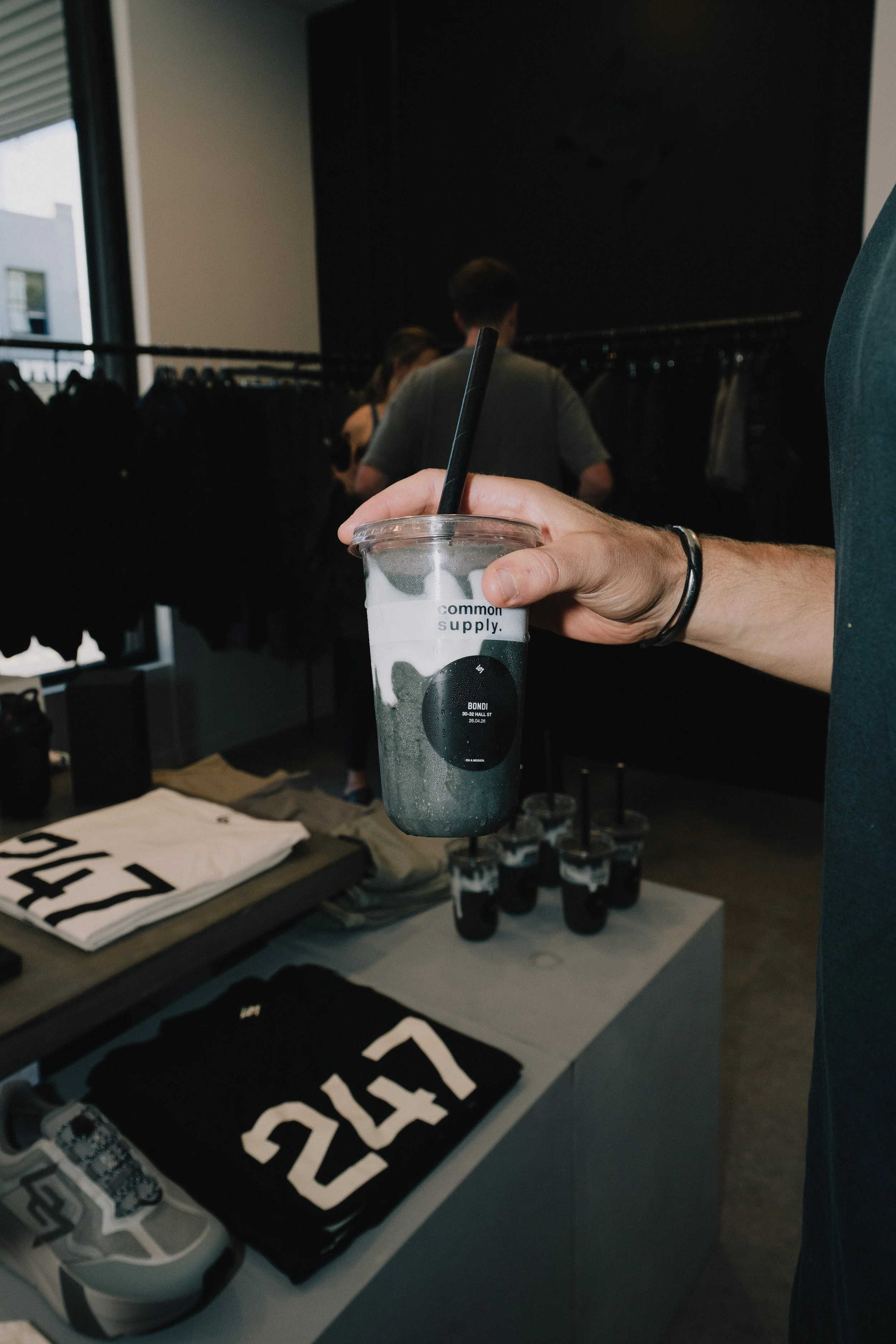 A person holding a clear plastic cup with a black straw, containing a dark-colored beverage, in a retail store. The store has clothing and accessories on display, including white and black items with bold text, and a shelving unit with multiple small