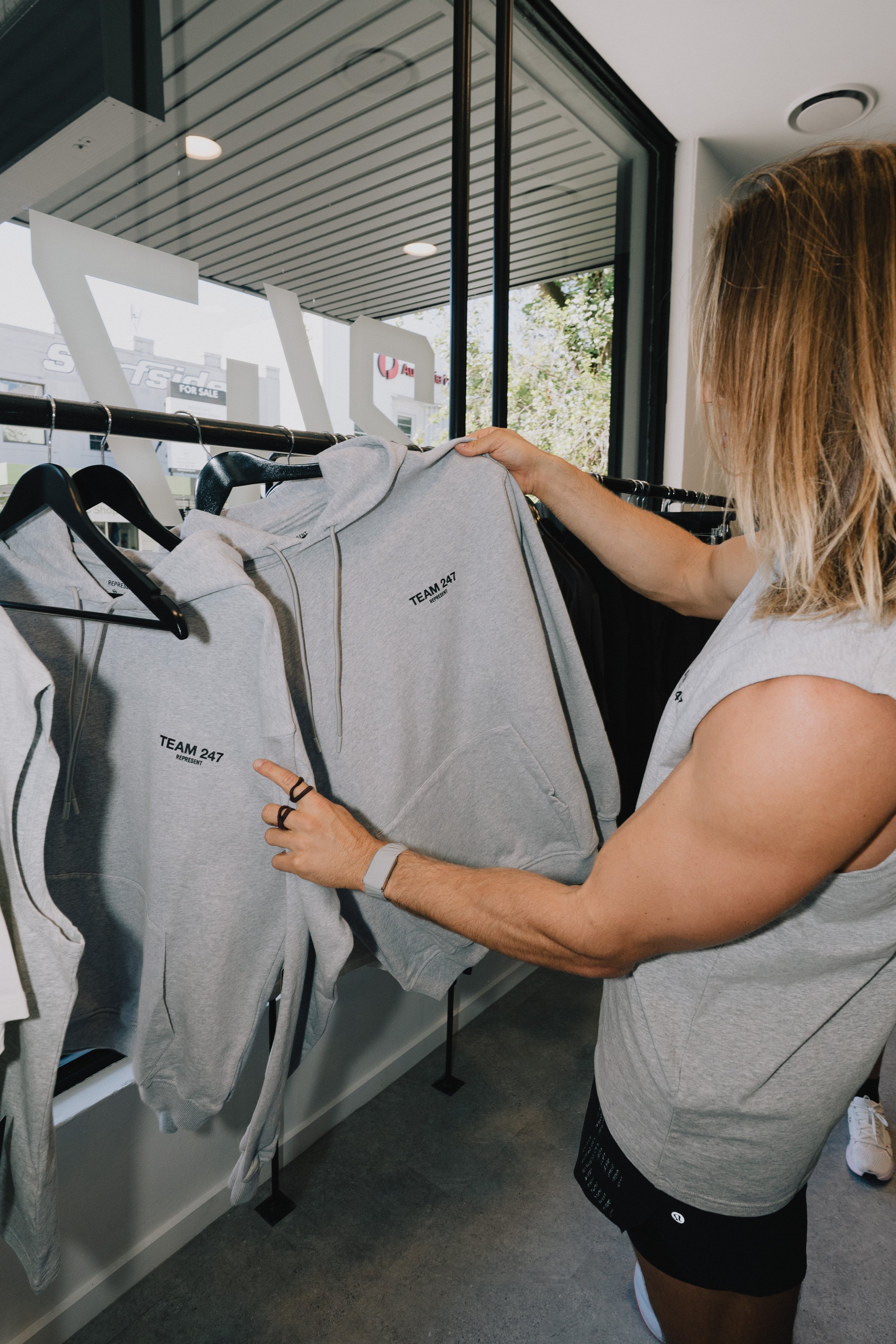 A person with blonde hair shopping in a store, examining grey hoodies with 'TEAM 247' printed on them, hanging on a black clothing rack near a window.