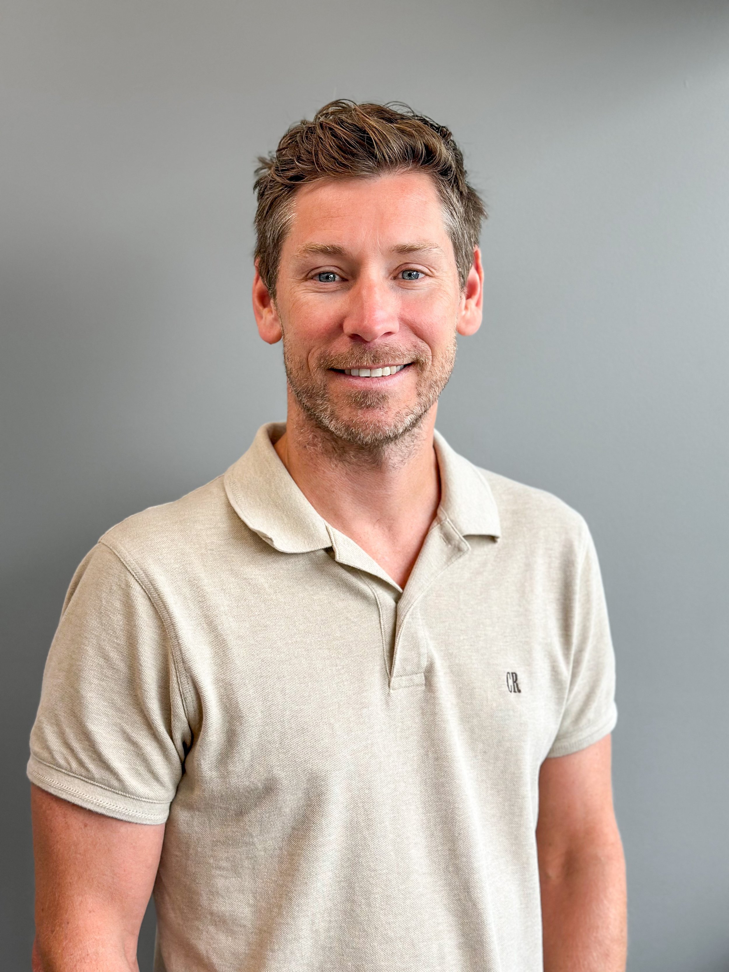 A man with short brown hair, blue eyes, and a light beard, smiling while wearing a beige collared shirt against a plain gray background.