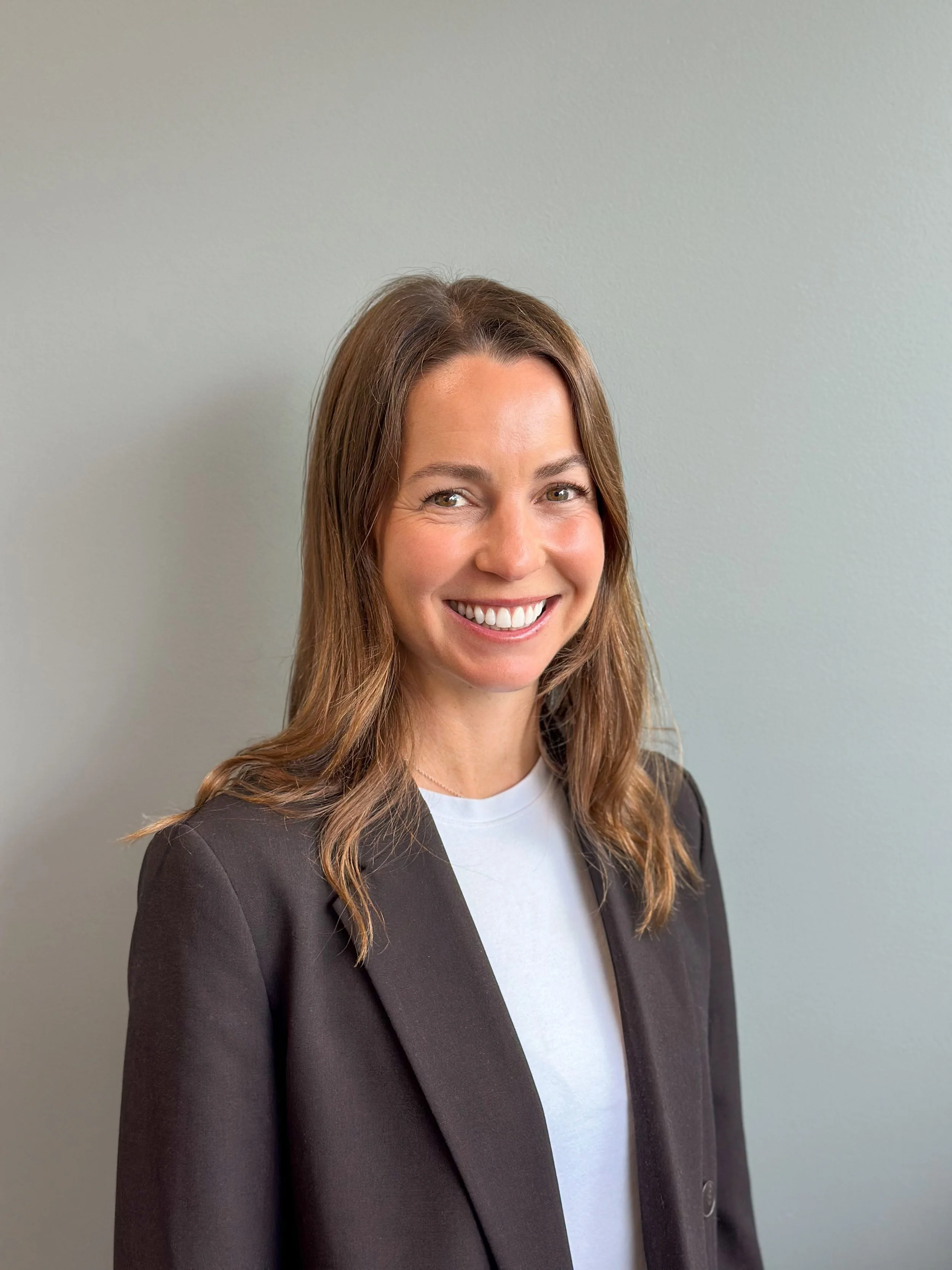 A smiling woman with light skin, long brown hair, wearing a white shirt and a black blazer, standing against a plain, light-colored wall.