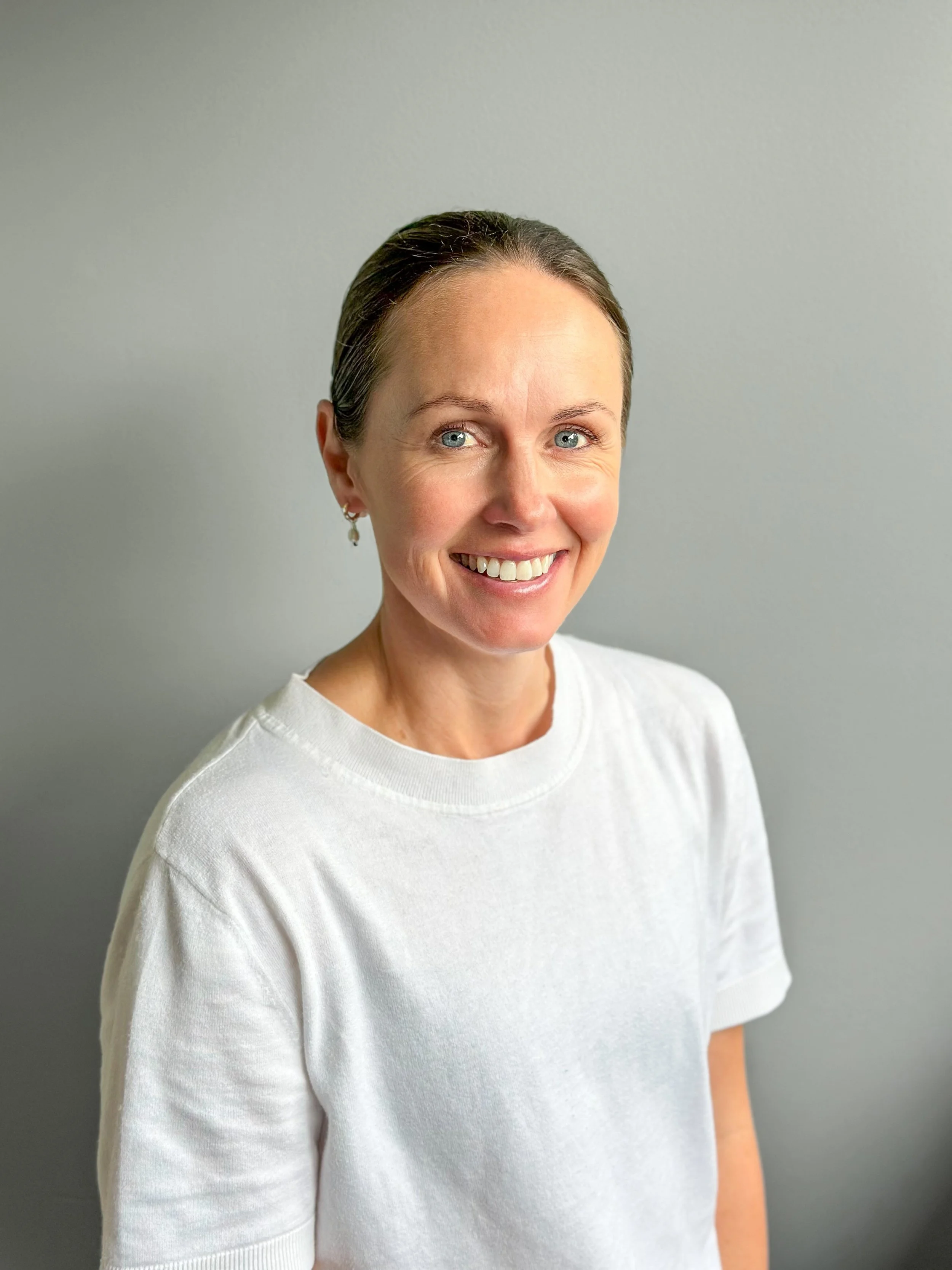 A smiling woman with short dark hair, wearing a white t-shirt, standing against a plain light gray background.