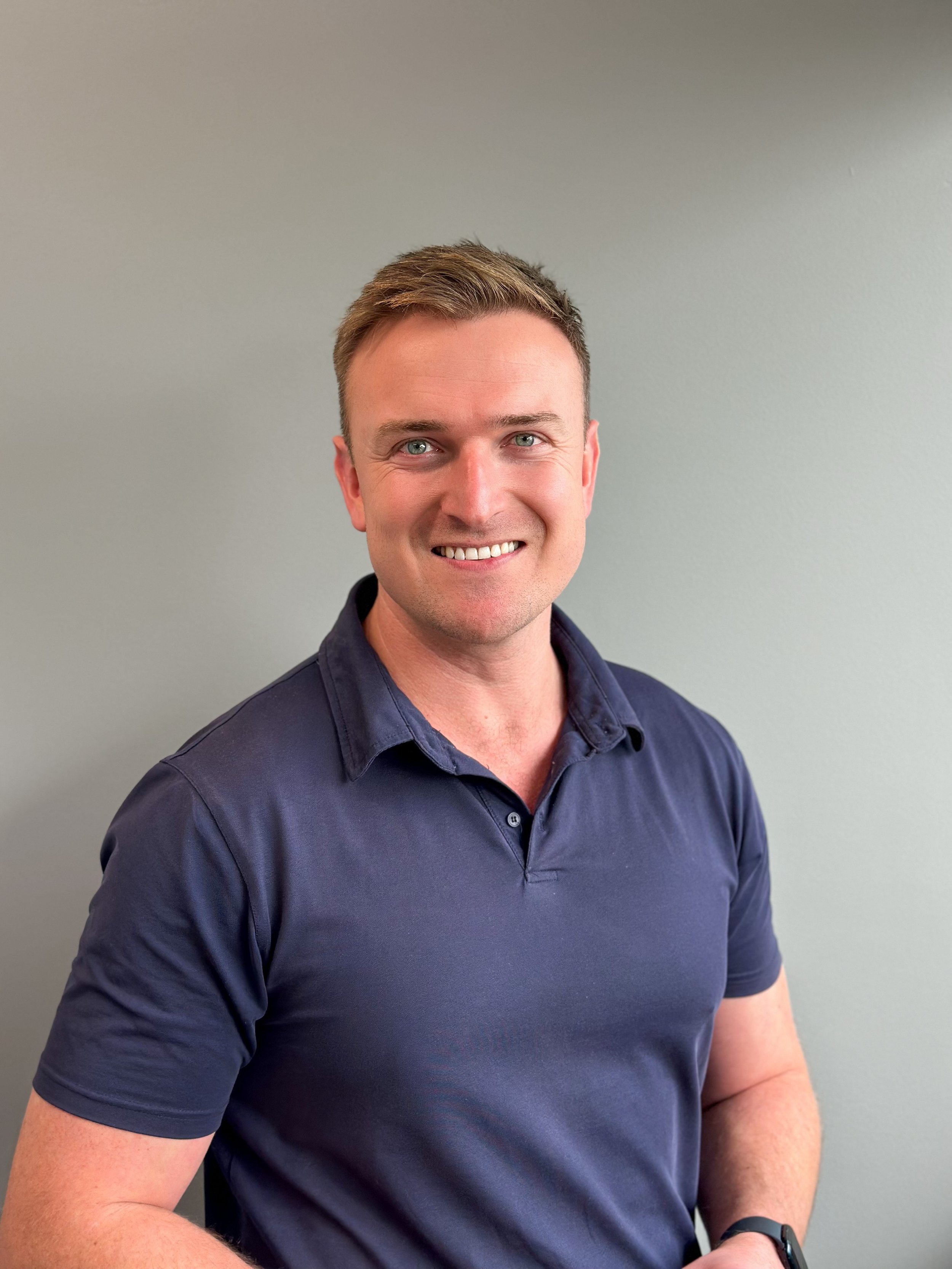 A man with light brown hair and blue eyes smiling, wearing a navy blue collared shirt, standing against a plain light gray wall.