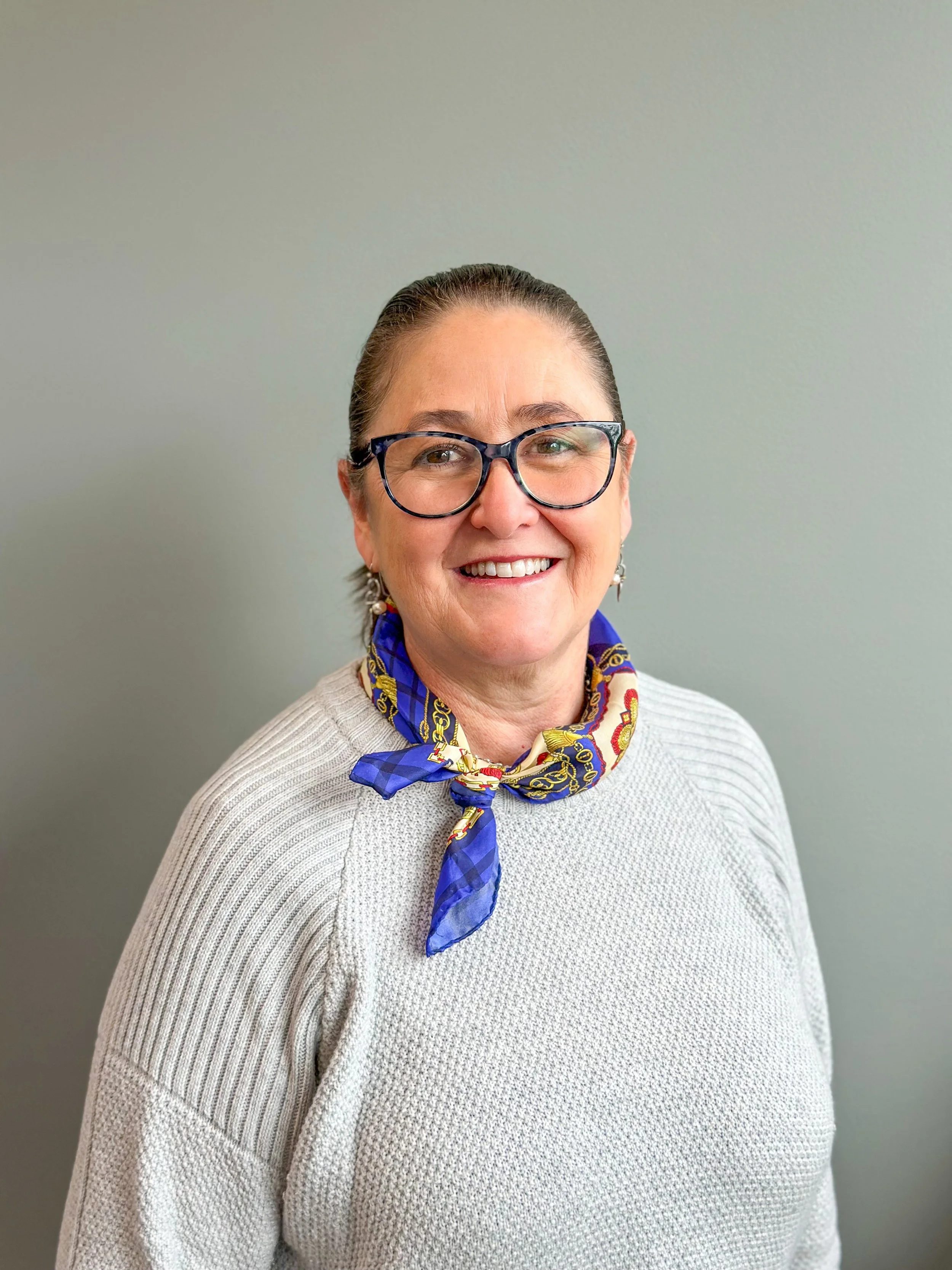 A middle-aged woman with glasses, short brown hair, smiling, wearing a white sweater and a blue patterned scarf, standing against a neutral background.