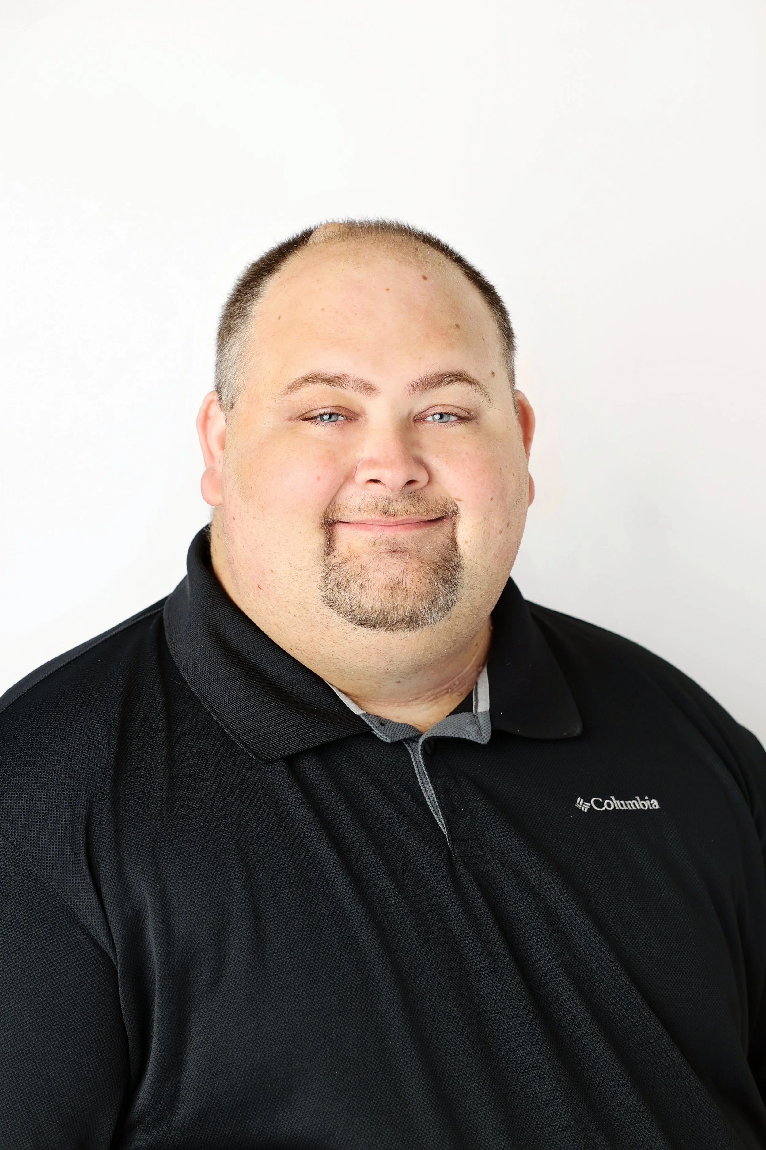 Portrait of a man with light skin and short dark hair, wearing a black Columbia jacket and a gray polo shirt, smiling slightly, set against a plain white background.