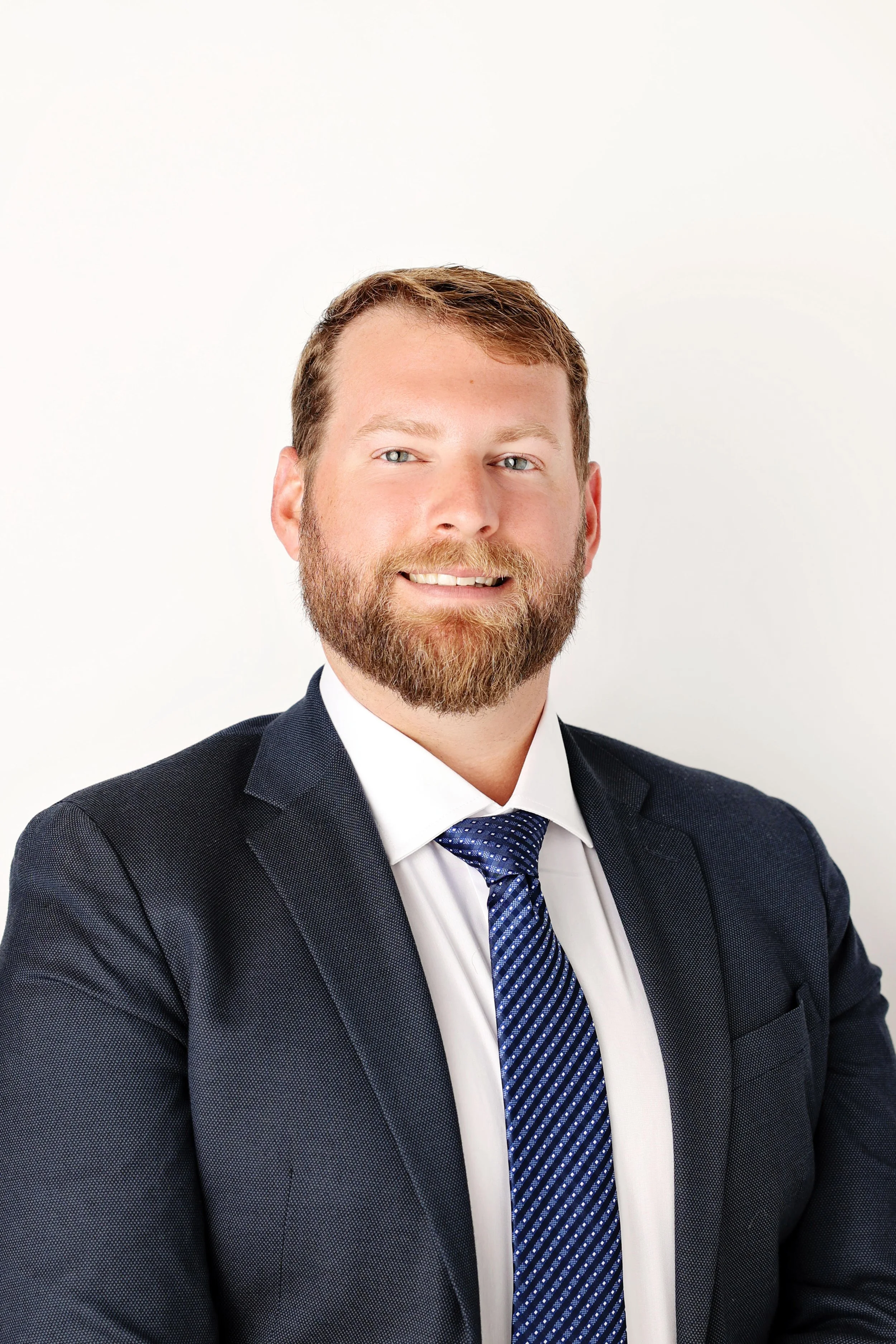 A man with a beard and short brown hair, dressed in a dark suit, white shirt, and a blue patterned tie, smiling at the camera against a plain white background.