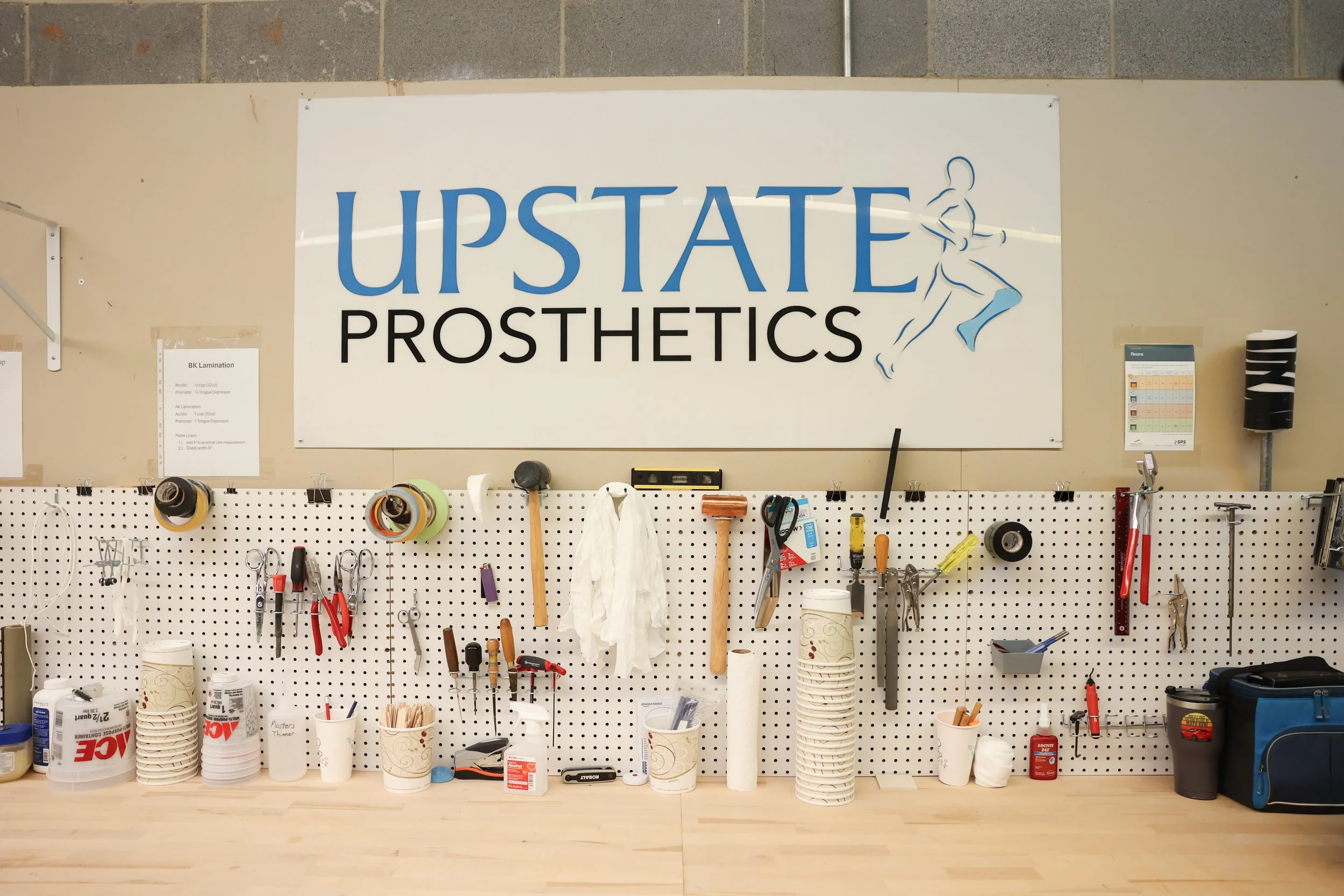 Workshop wall with tools and supplies, including scissors, hammers, and tape, under a sign that reads 'Upstate Prosthetics'.