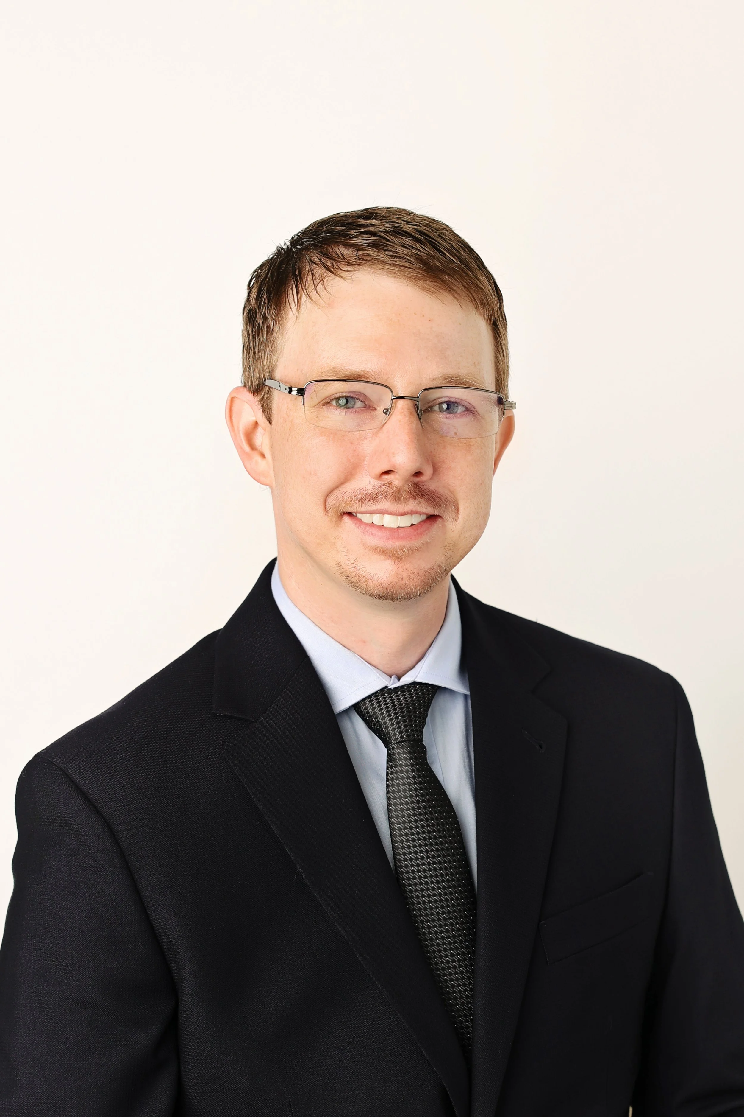 Headshot of a young man with short brown hair, wearing glasses, a black suit, blue shirt, and patterned tie, smiling against a plain light background.