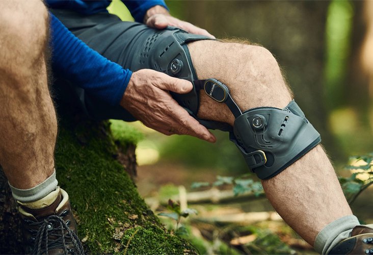 A person with a prosthetic leg resting outdoors in a forest, with a hand adjusting the prosthetic.