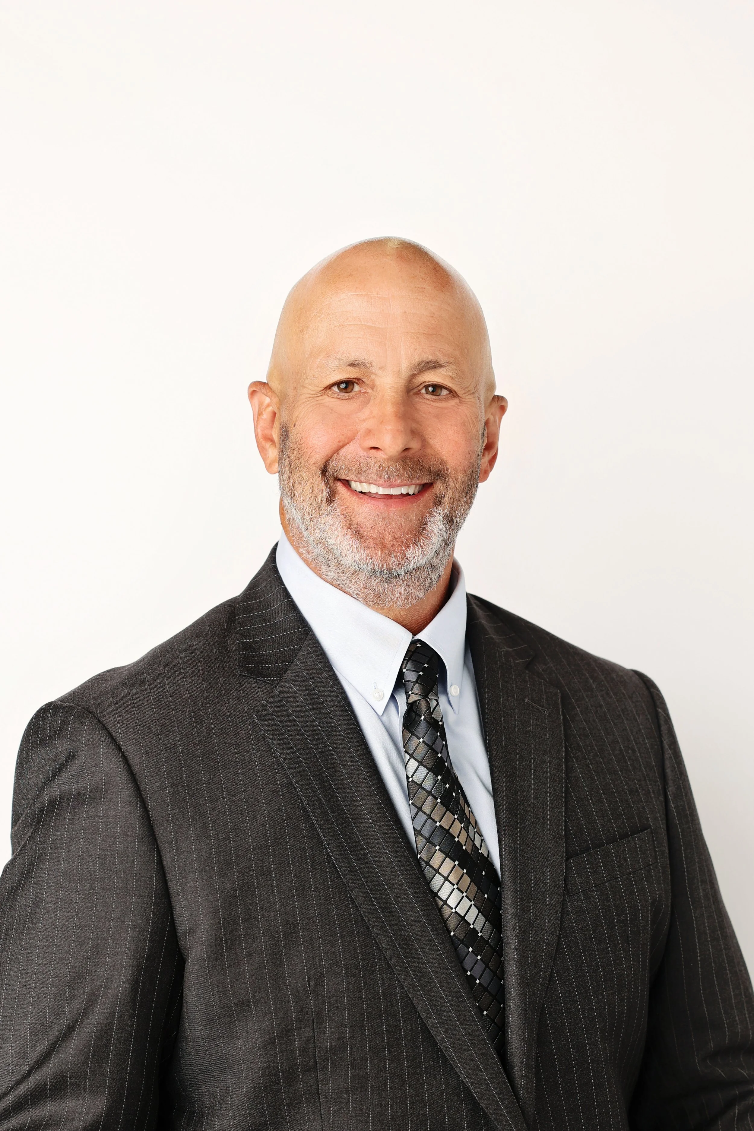 A middle-aged man with a shaved head, beard, and mustache, wearing a dark gray pinstripe suit, white shirt, and a patterned tie, smiling against a plain white background.