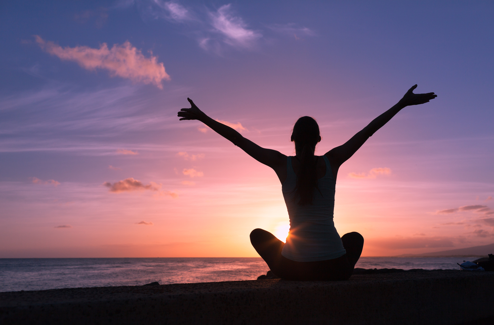 Person sitting cross-legged on a beach at sunset with arms raised in a meditation pose.