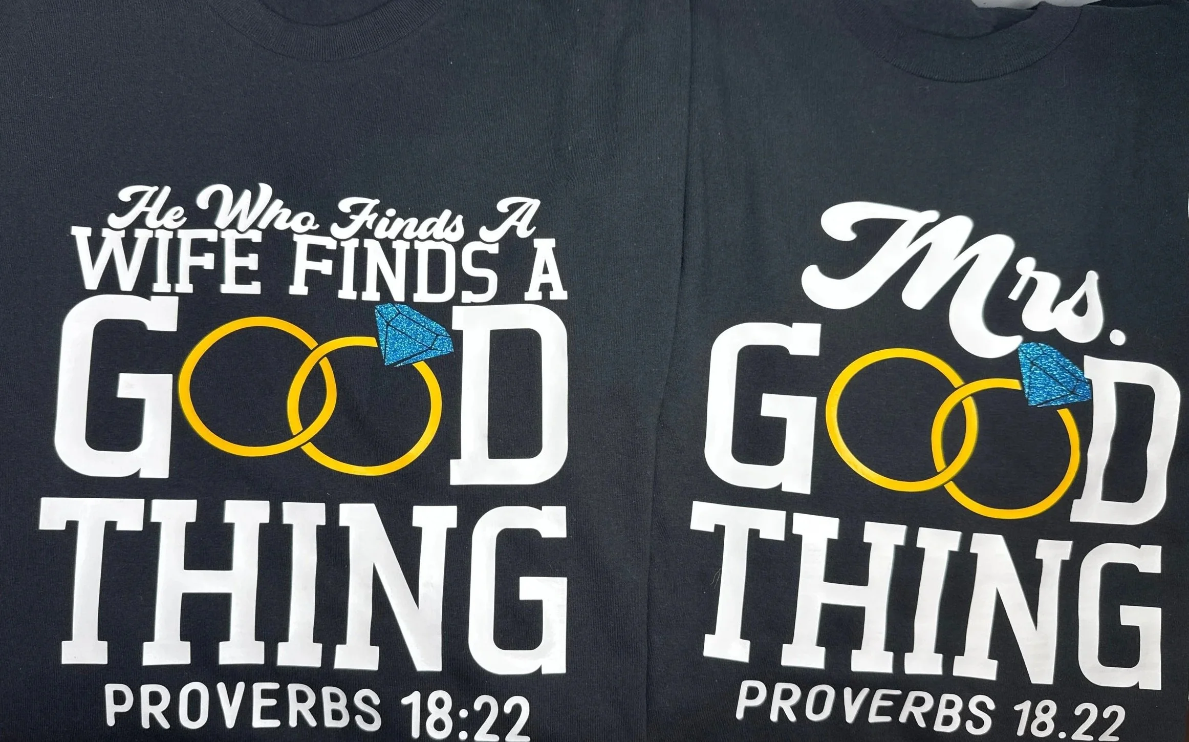 Dark-colored T-shirts with printed Christian-themed text in white and yellow, including a blue diamond and yellow rings, referencing Proverbs 18:22.