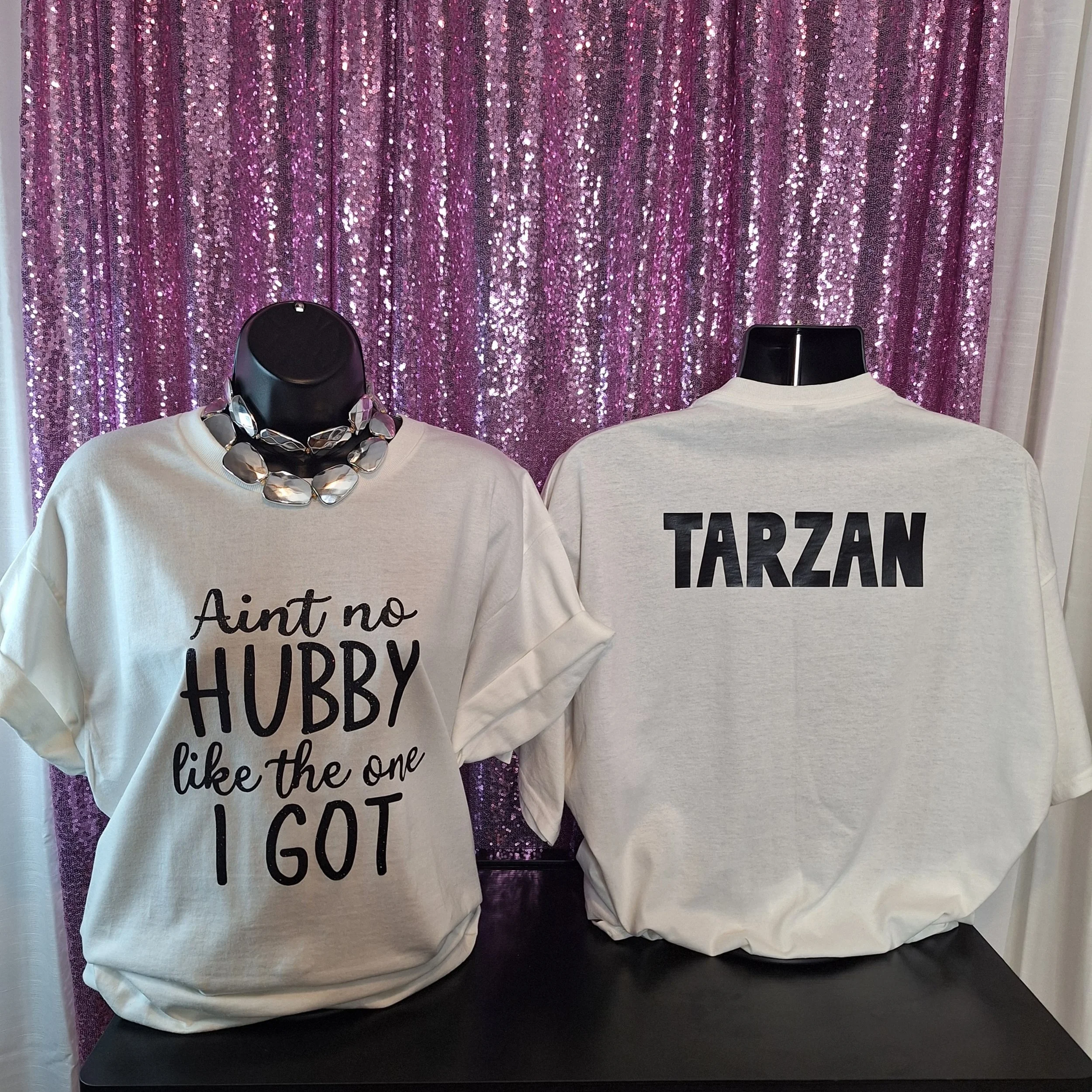 Two mannequins dressed in casual clothing placed in front of a purple, shiny, sequined curtain backdrop. The mannequin on the left wears a white top with black words and a statement necklace. The mannequin on the right wears a gray top with black text. Part of a third mannequin is visible on the right side, also in white. The mannequins are positioned on a black surface.